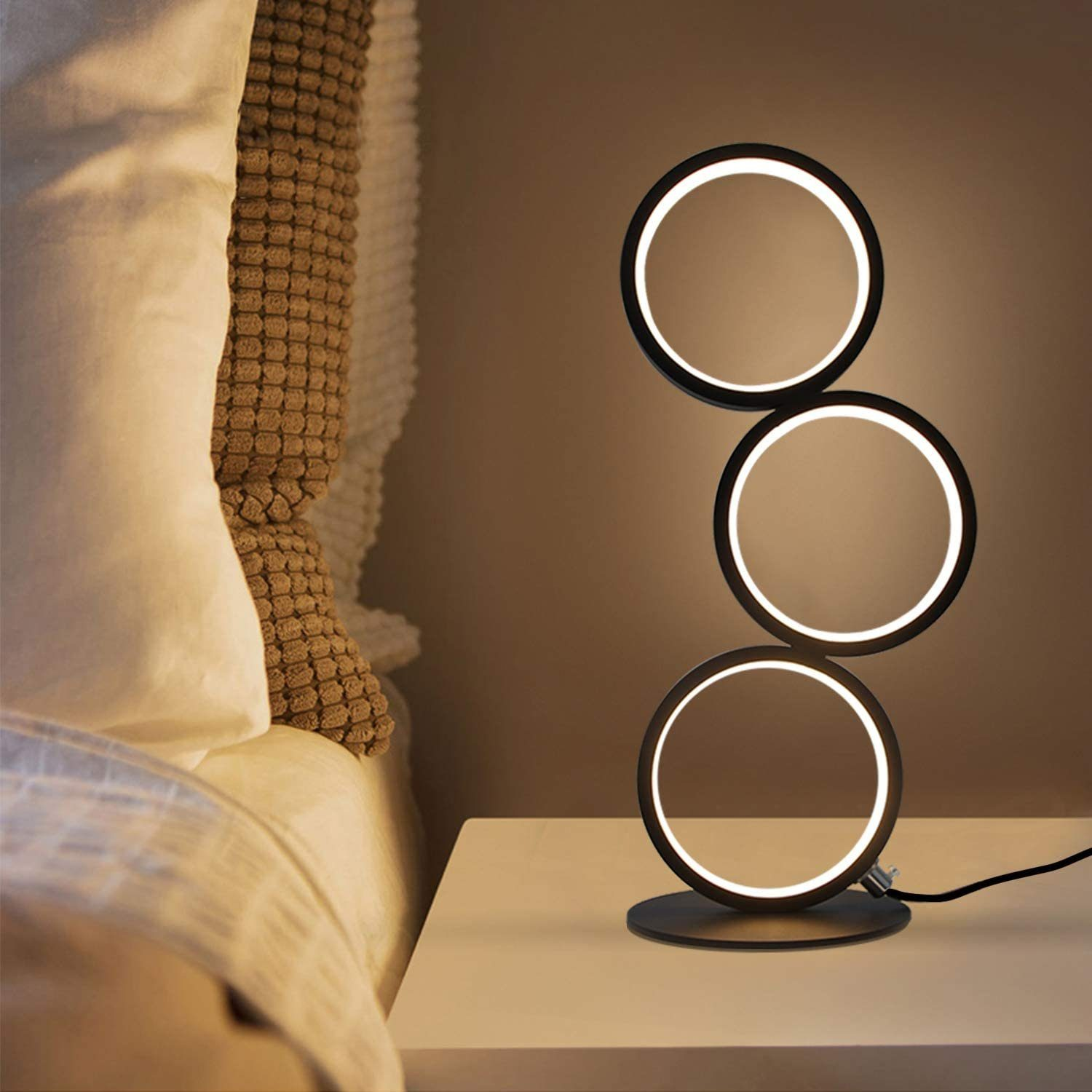 Una lampada moderna con tre anelli neri illuminati su un comodino. Un letto con cuscini e una coperta sullo sfondo.
