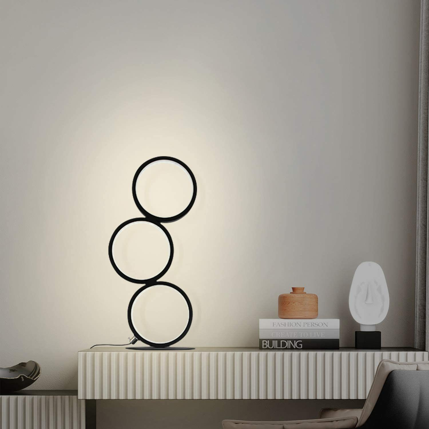 Una lampada moderna con tre luci circolari collegate. Accanto, un tavolo con libri, una scultura e un contenitore di legno. Sfondo neutro.