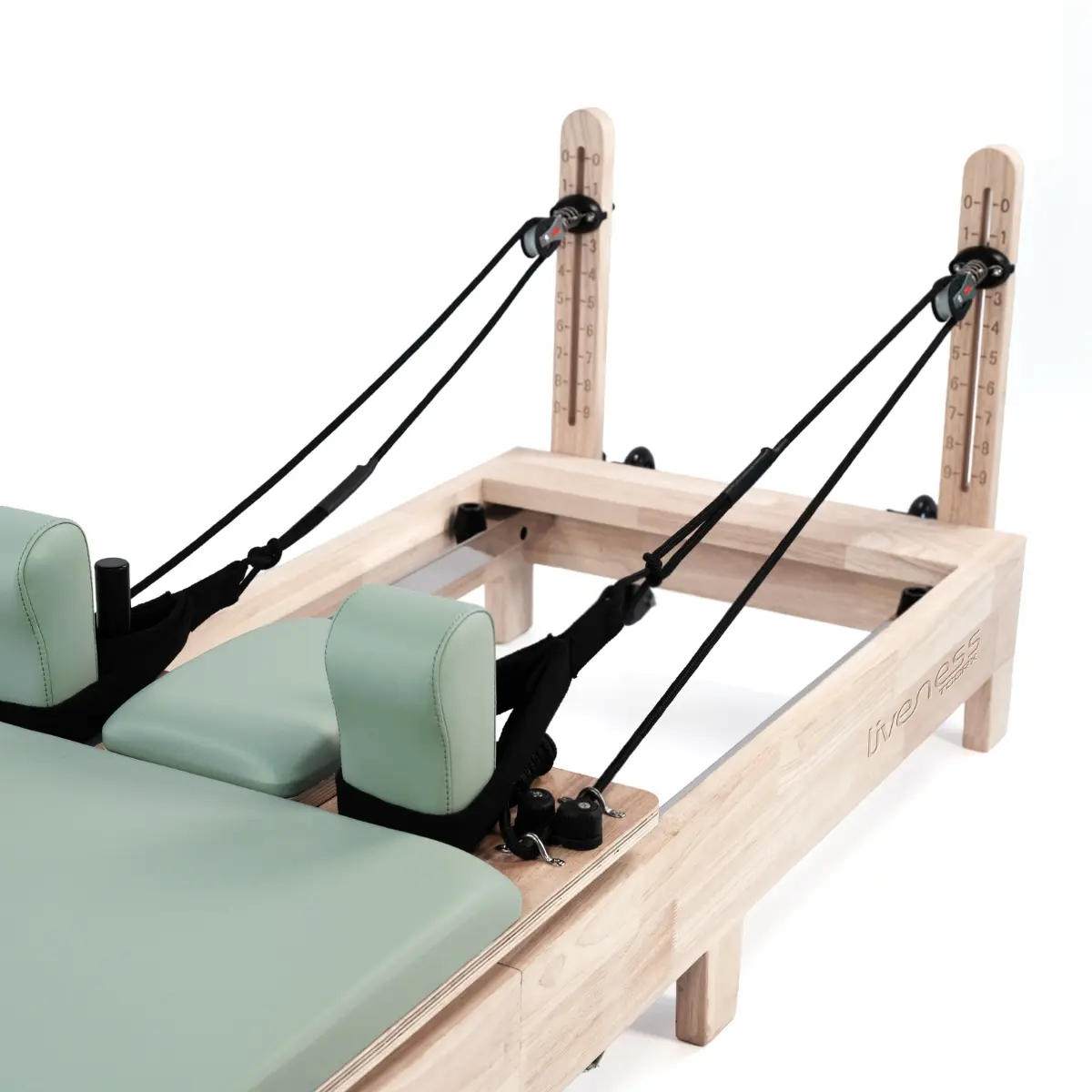 Pilates reformer machine. Licht houten frame, lichtgroene vulling en hoofdsteunen. Zwarte riemen en katrollen. Witte achtergrond.
