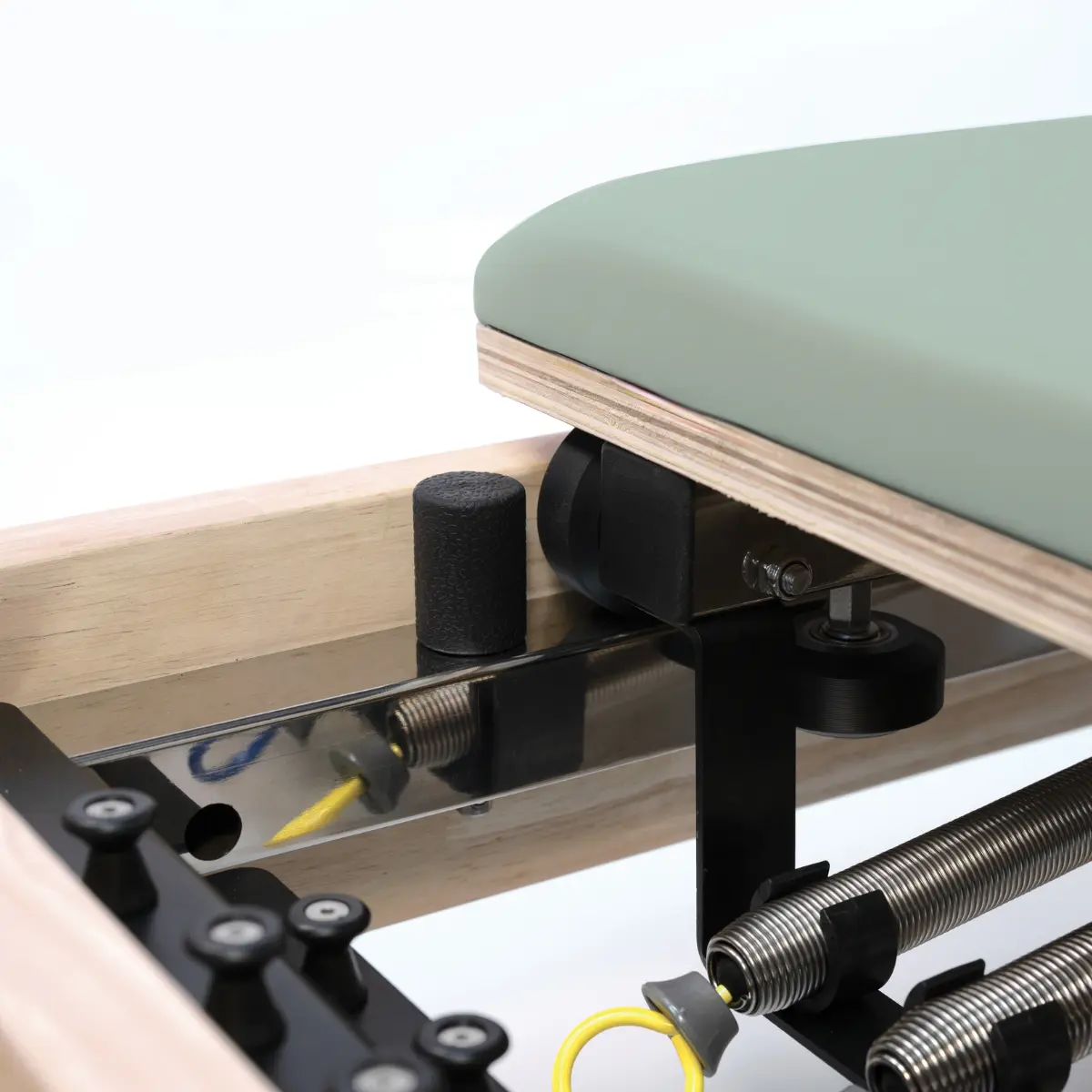 Een Pilates-machine met een groen kussen en metalen veren. Er is een houten frame en zwarte metalen componenten. De achtergrond is wit.