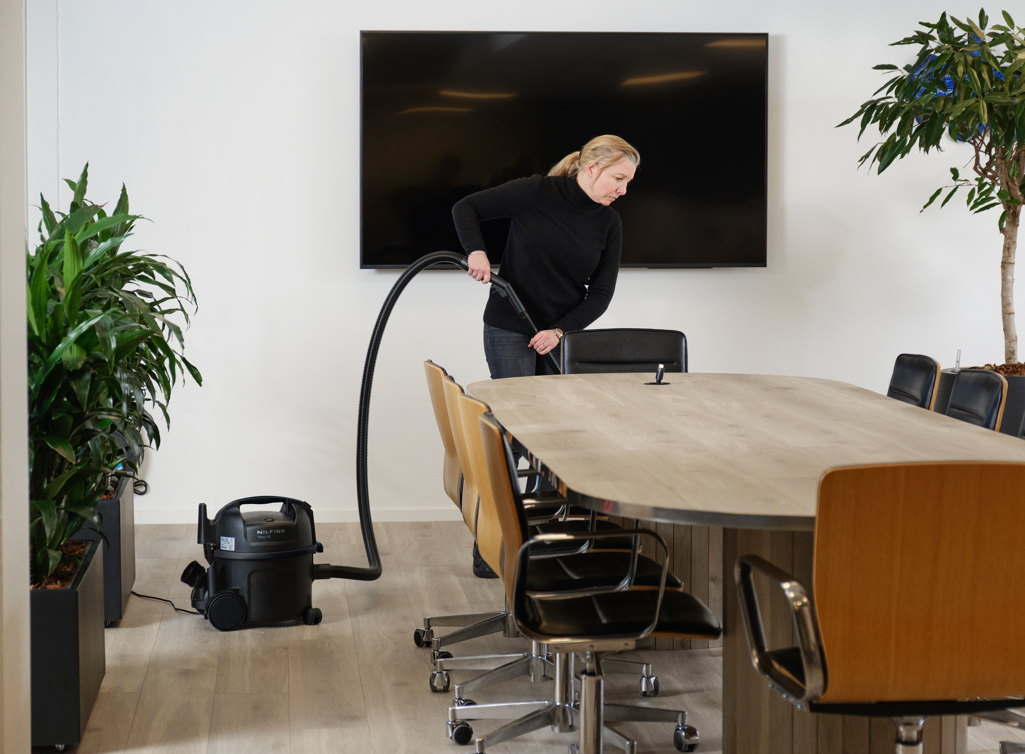 Een vrouw in het zwart gebruikt een stofzuiger in een moderne vergaderruimte. Een groot scherm en een tafel met stoelen zijn zichtbaar.