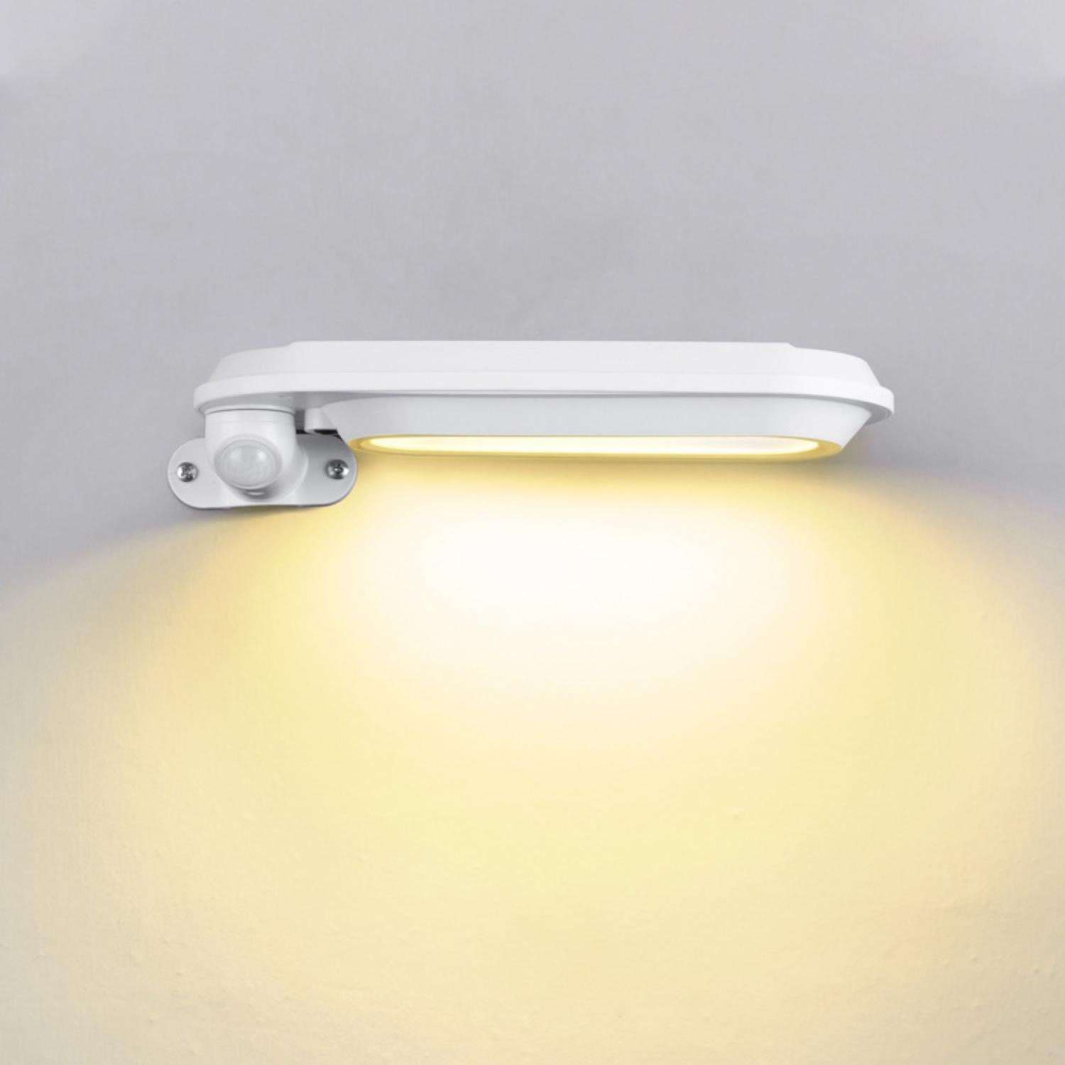Eine weiße LED-Wandleuchte mit Bewegungssensor ist an einer hellgrauen Wand montiert. Das Licht ist an und beleuchtet die Wand darunter.