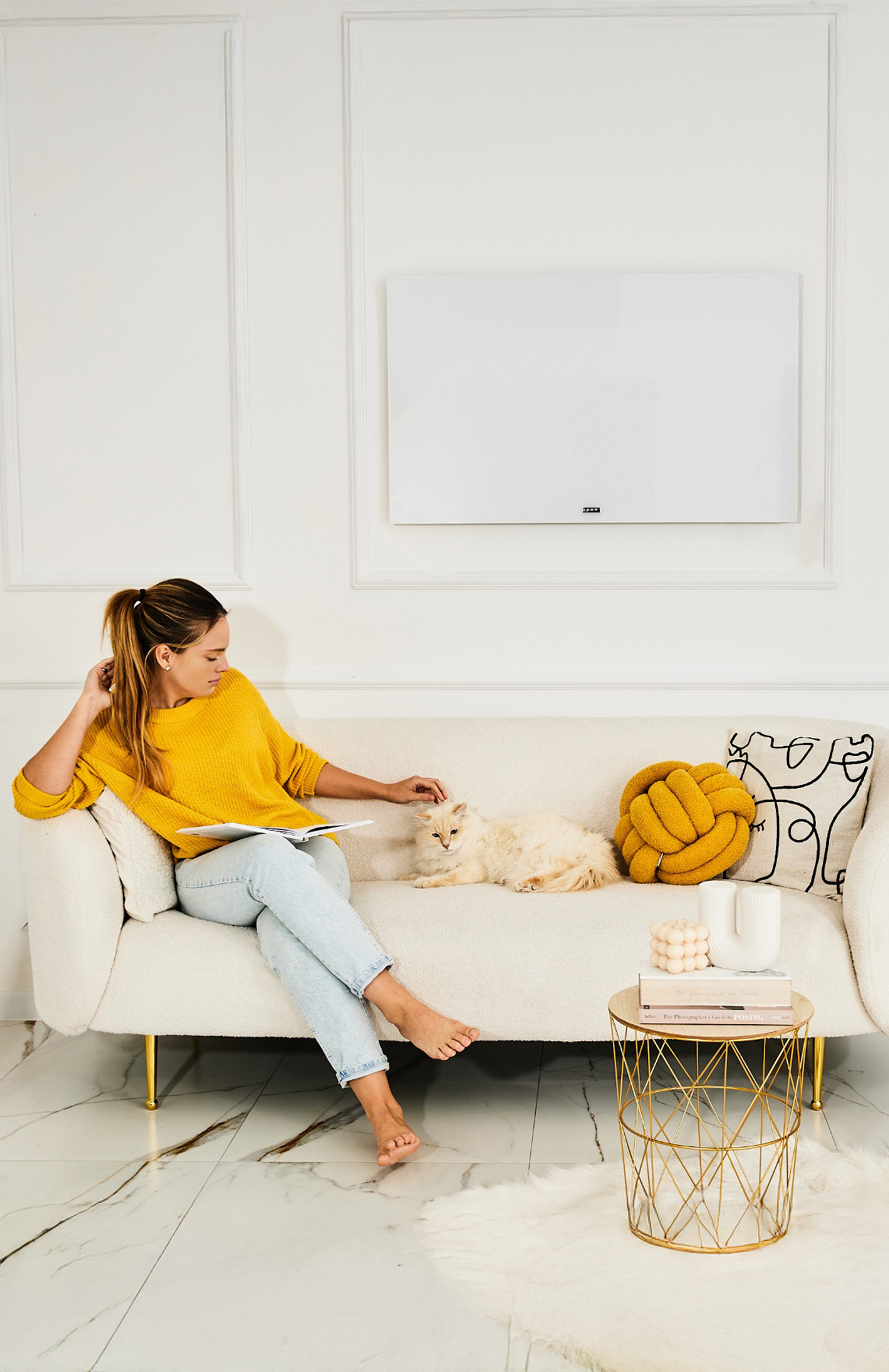 Kobieta w żółtym swetrze głaszcze kota na białej sofie. Po prawej stronie złoty stolik.