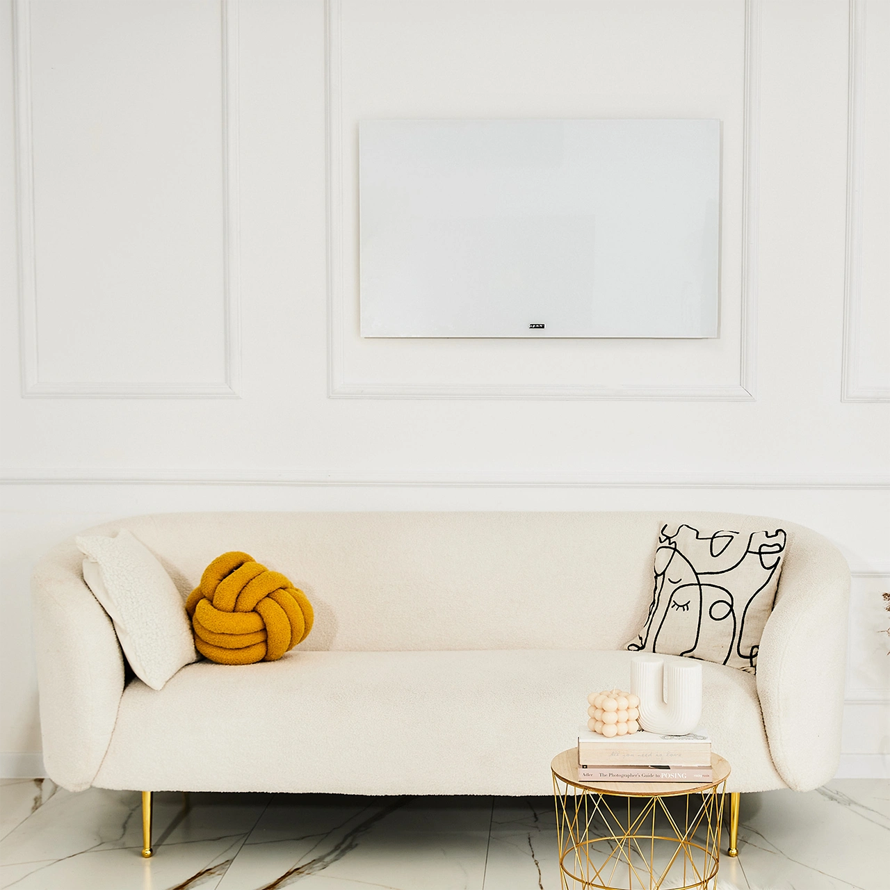 Biała sofa z poduszkami, poduszką w kształcie węzła i stolikiem z świecą i książkami. Biały panel na ścianie.