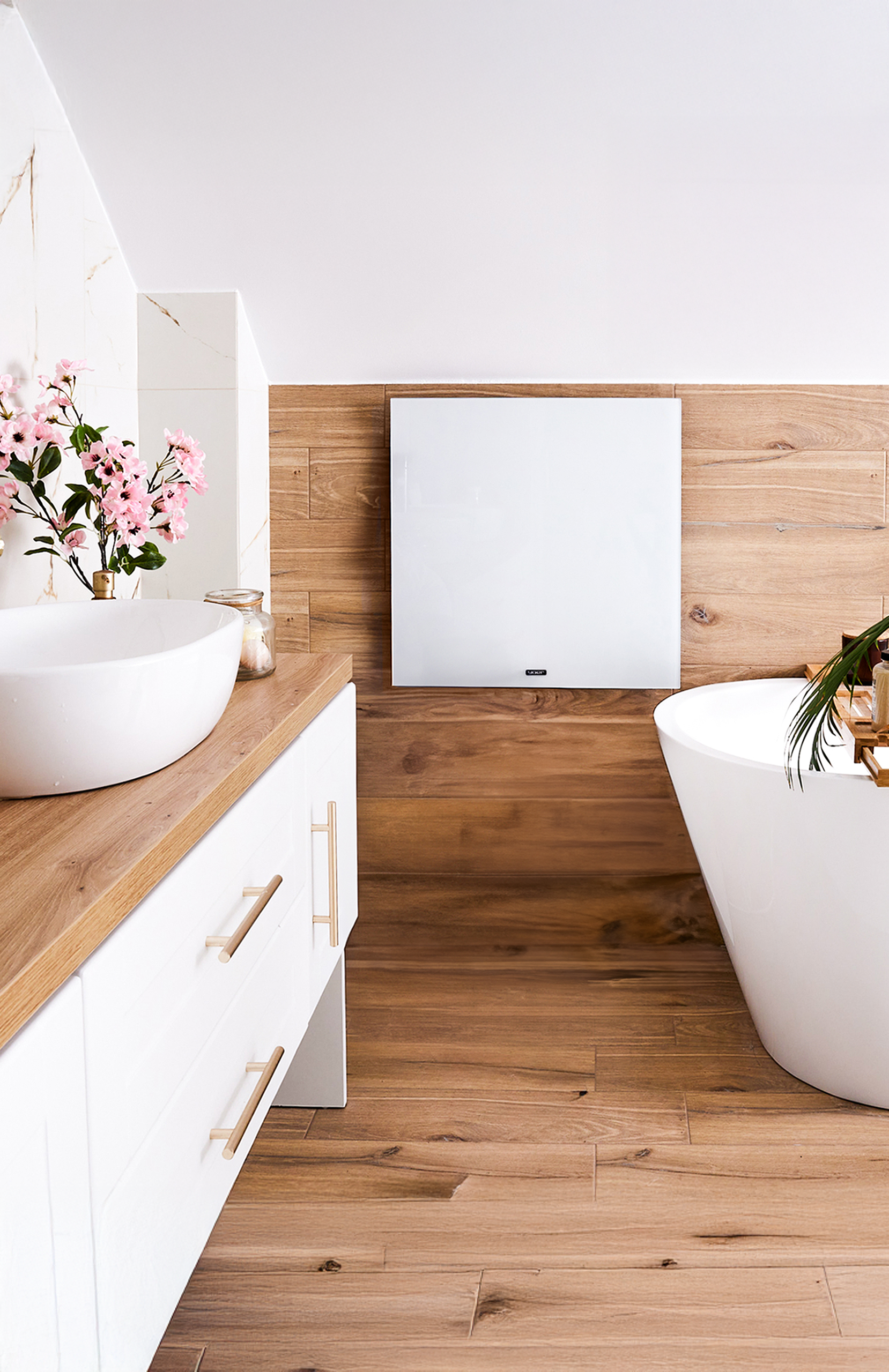 Nowoczesna łazienka z białą umywalką, szafkami i wanną. Drewniane akcenty i różowe kwiaty zdobią przestrzeń.