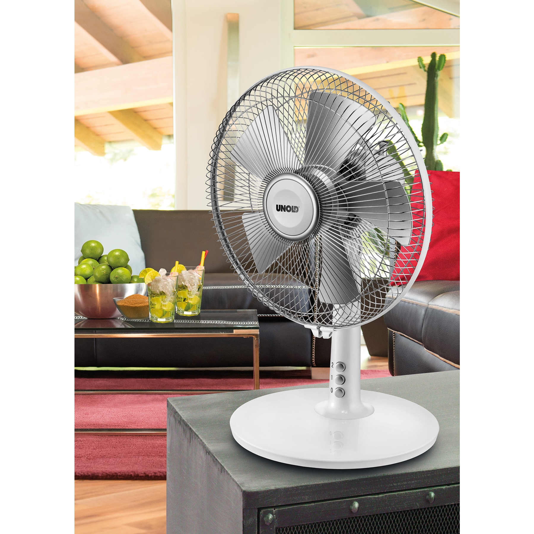 Ein weißer Unold-Ventilator steht auf einem grauen Tisch. Cocktails und Limetten stehen auf einem Couchtisch. Couch und Kaktus sichtbar.