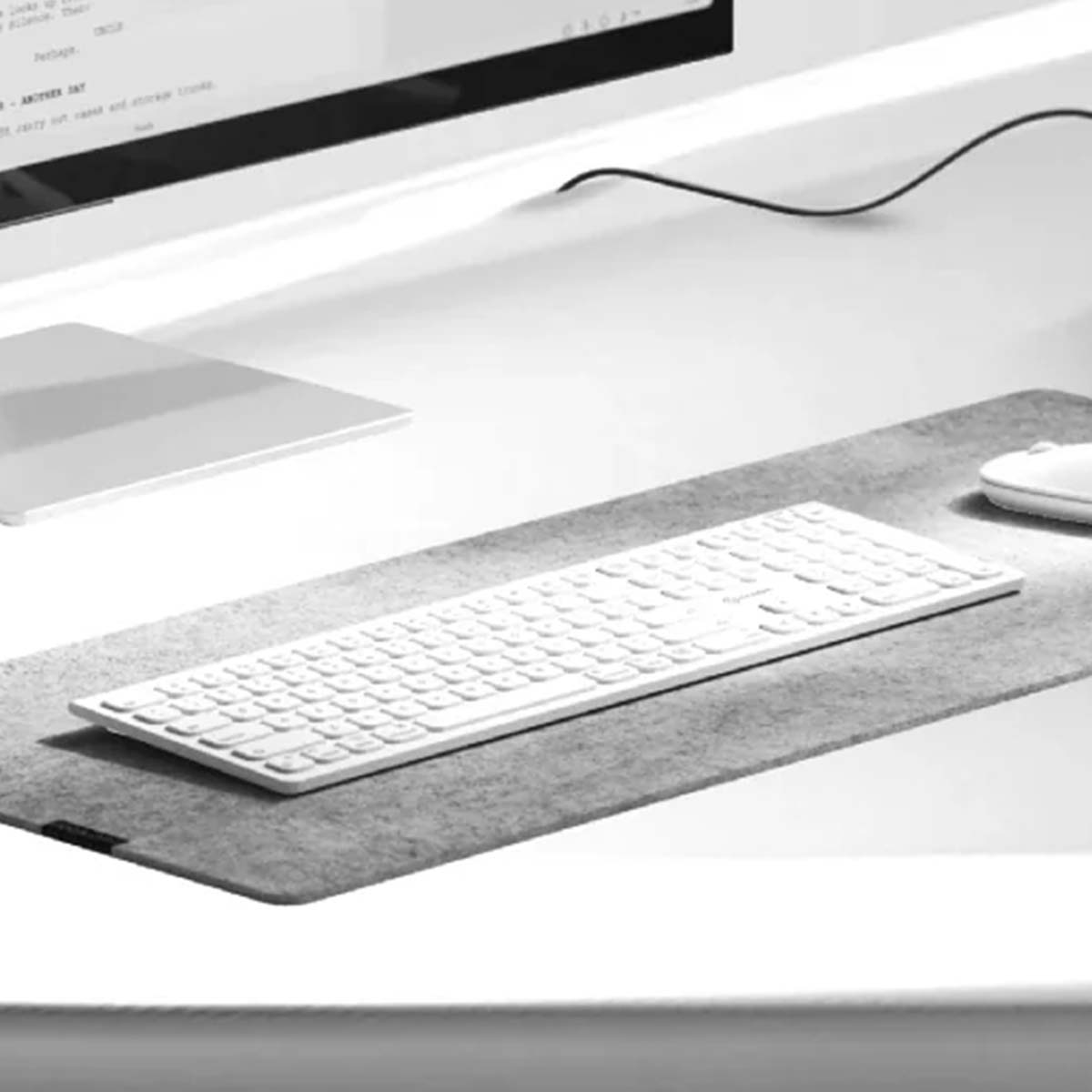 Una tastiera e un mouse bianchi su un tappetino grigio. Sullo sfondo uno schermo del computer. La scena è in scala di grigi.