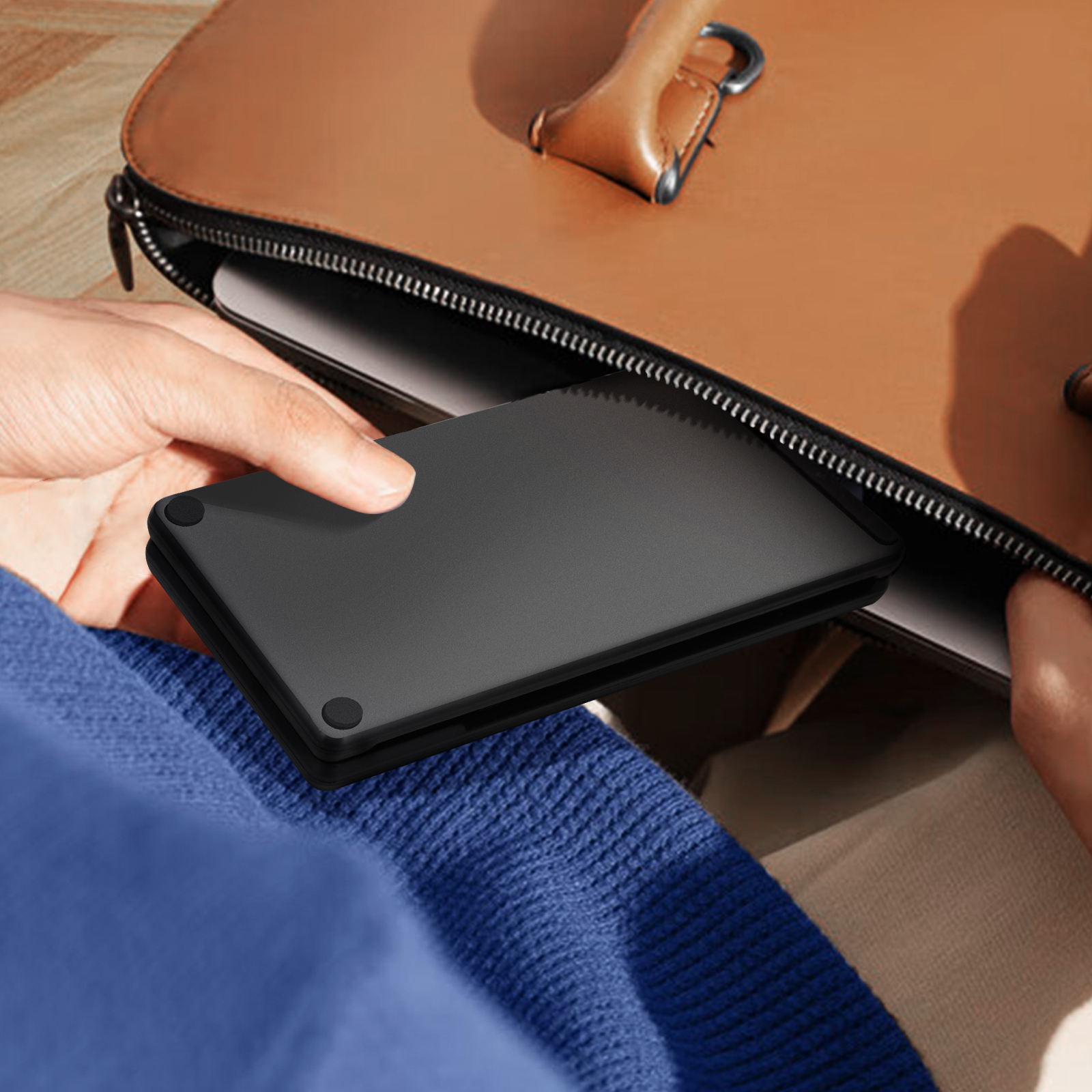 Una persona tiene in mano una custodia nera per laptop e la mette in una borsa di pelle marrone. La persona indossa un maglione blu.