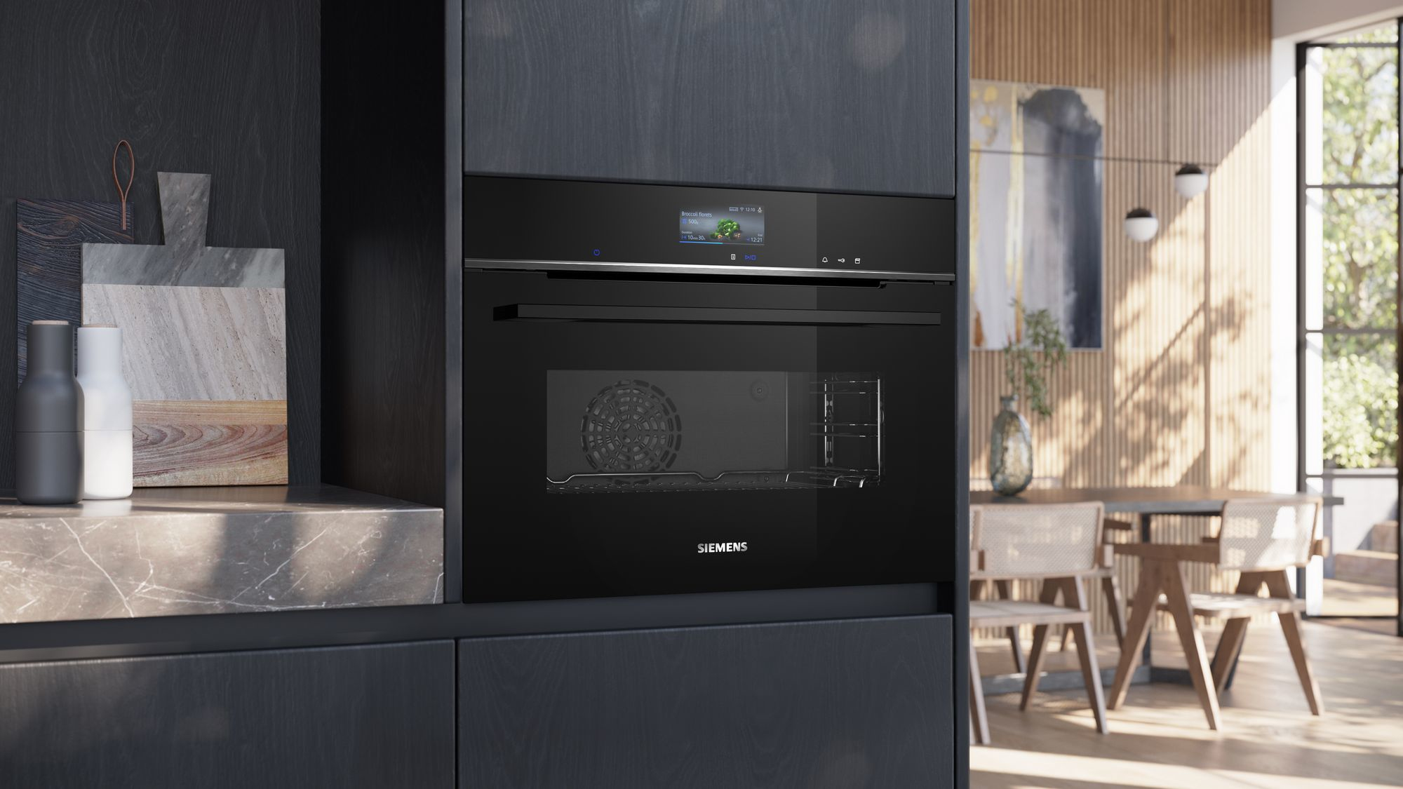 Een zwarte Siemens oven is ingebouwd in een keuken. Een scherm toont kookinfo. Snijplanken en een eethoek zijn ook zichtbaar.