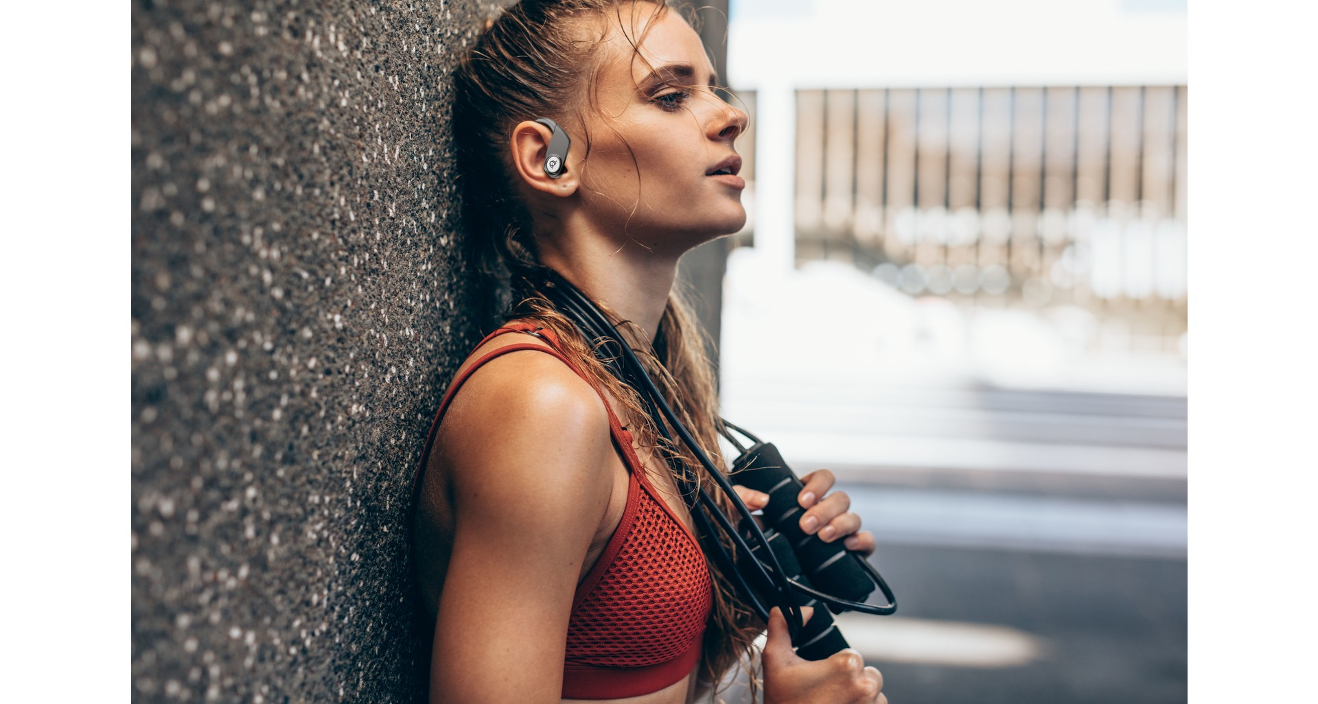 Eine Frau lehnt an einer Wand und hält ein Springseil. Sie trägt einen roten Sport-BH und hat einen Ohrhörer im Ohr. Der Hintergrund ist verschwommen.