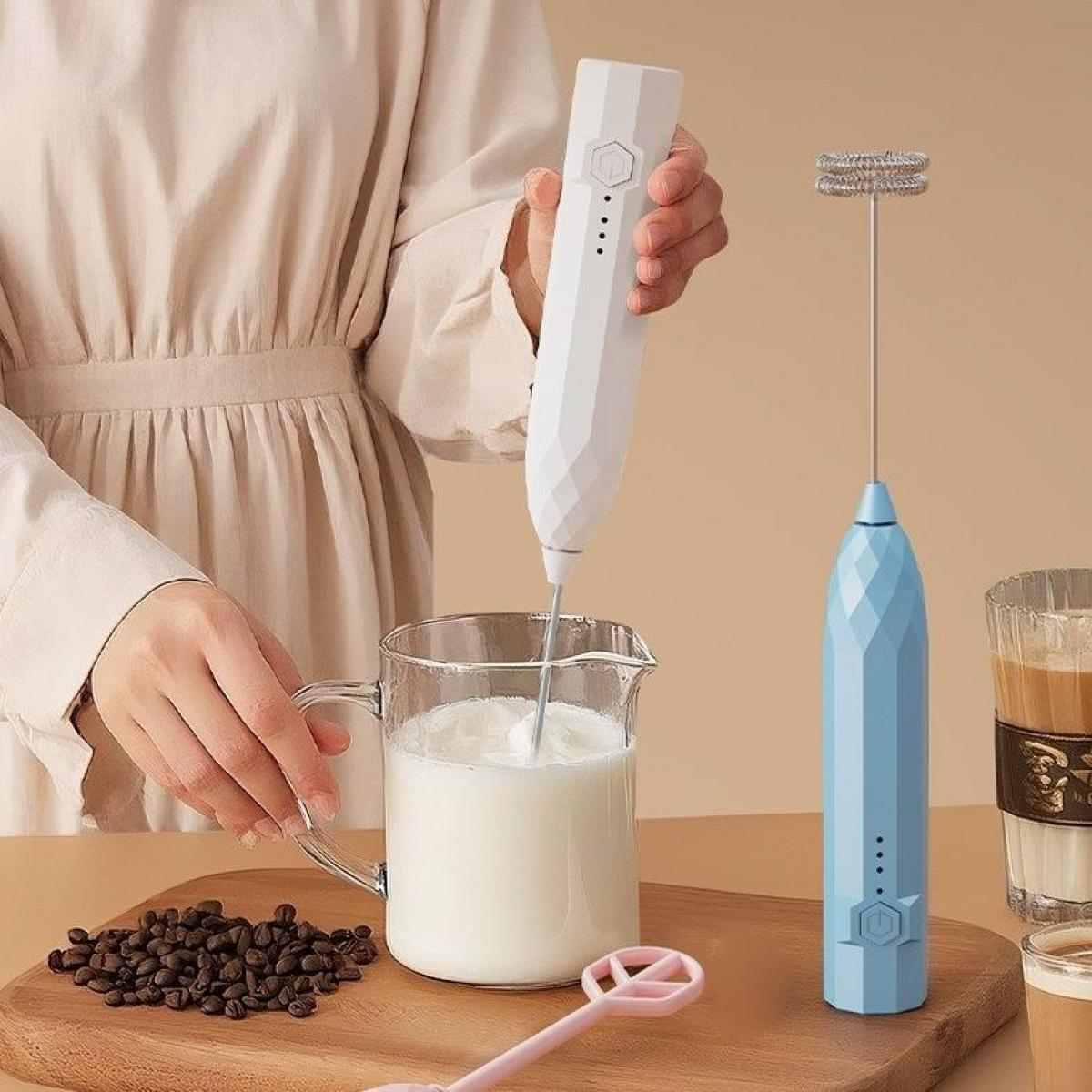 Eine Person schäumt Milch mit einem weißen Aufschäumer auf. Ein blauer Aufschäumer steht daneben. Kaffeebohnen und ein rosa Löffel liegen auf Holz.