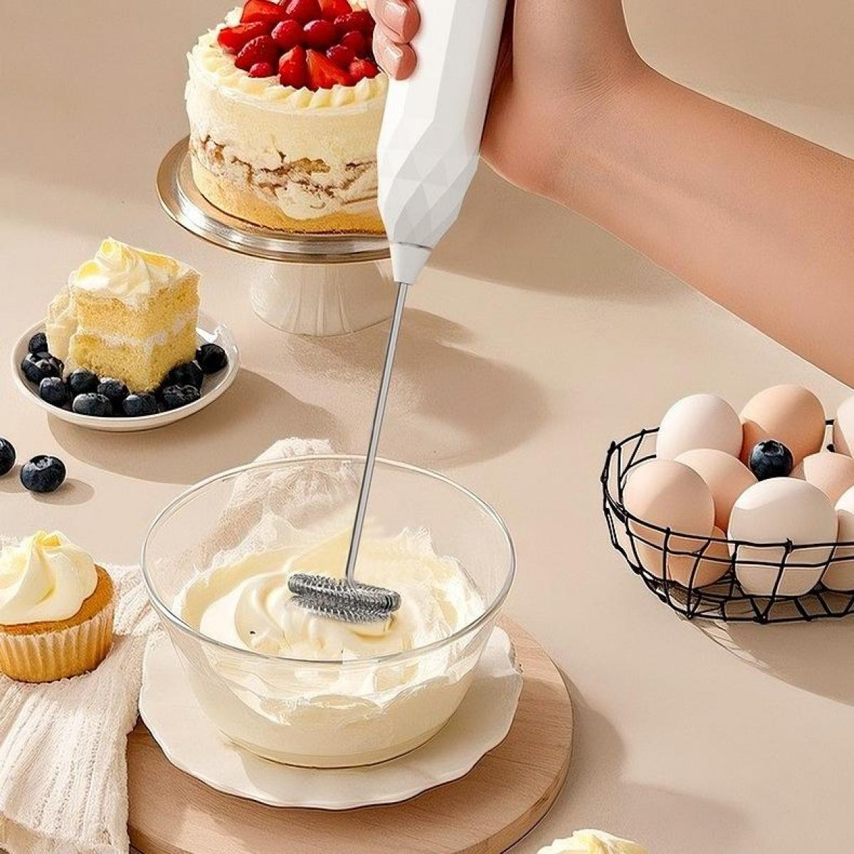 Eine Hand hält einen Milchaufschäumer über einer Schüssel Sahne. Ein Kuchen mit Erdbeeren, Kuchenstück mit Blaubeeren, Eier und ein Cupcake sind auf dem Tisch.
