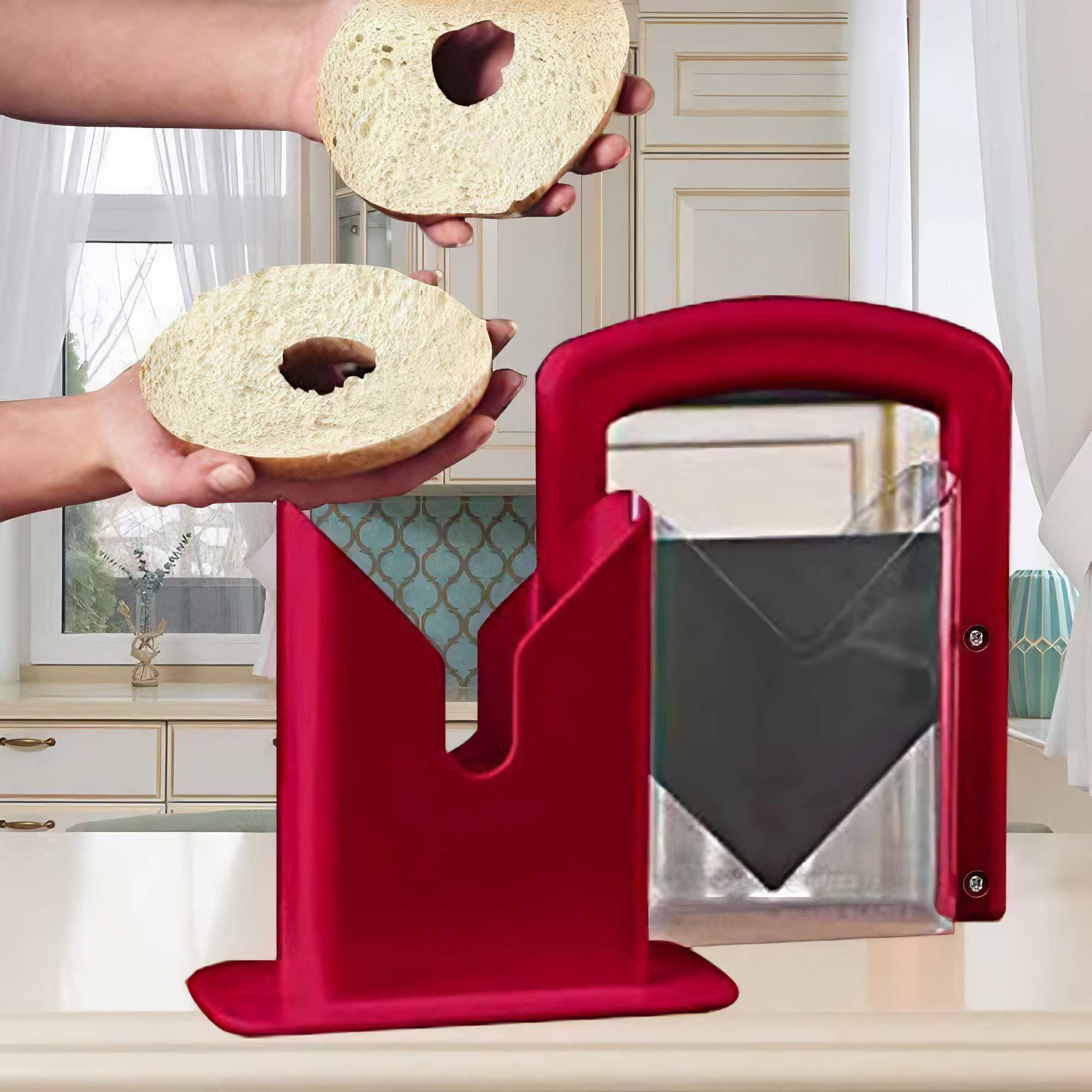 Hände halten Bagel-Hälften. Vorne ein roter Bagel-Schneider. Küchenhintergrund mit Schränken und Fenstern.