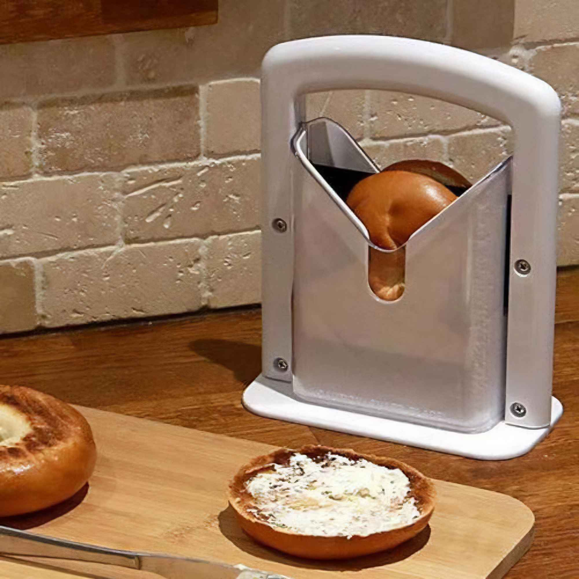Ein Bagel-Schneider mit einem Bagel darin. Ein geschnittener Bagel mit Frischkäse und ein Messer liegen auf einem Holzbrett.