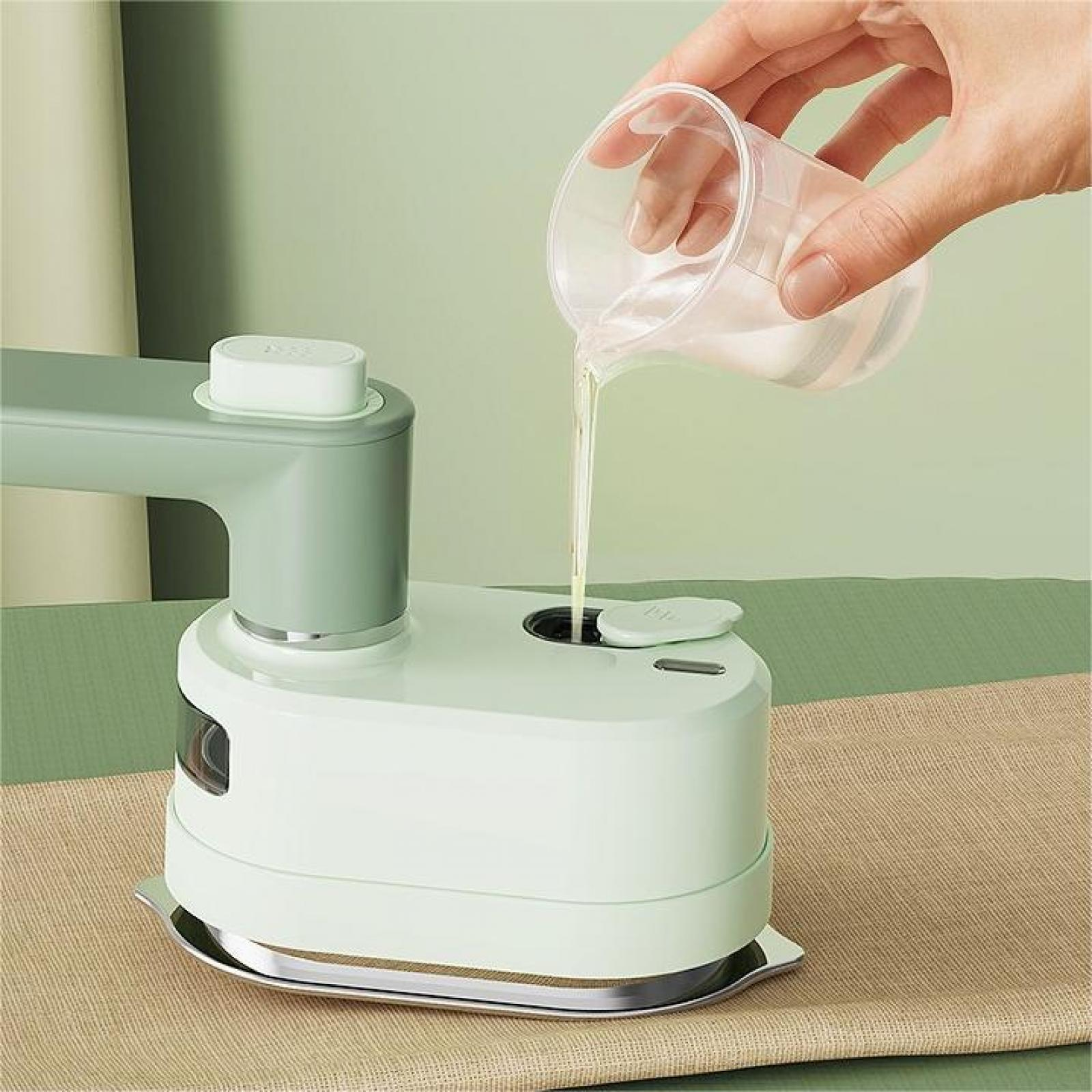 Eine Hand gießt Flüssigkeit aus einem durchsichtigen Becher in ein weiß-grünes Gerät auf einer braunen Oberfläche, grüner Hintergrund.