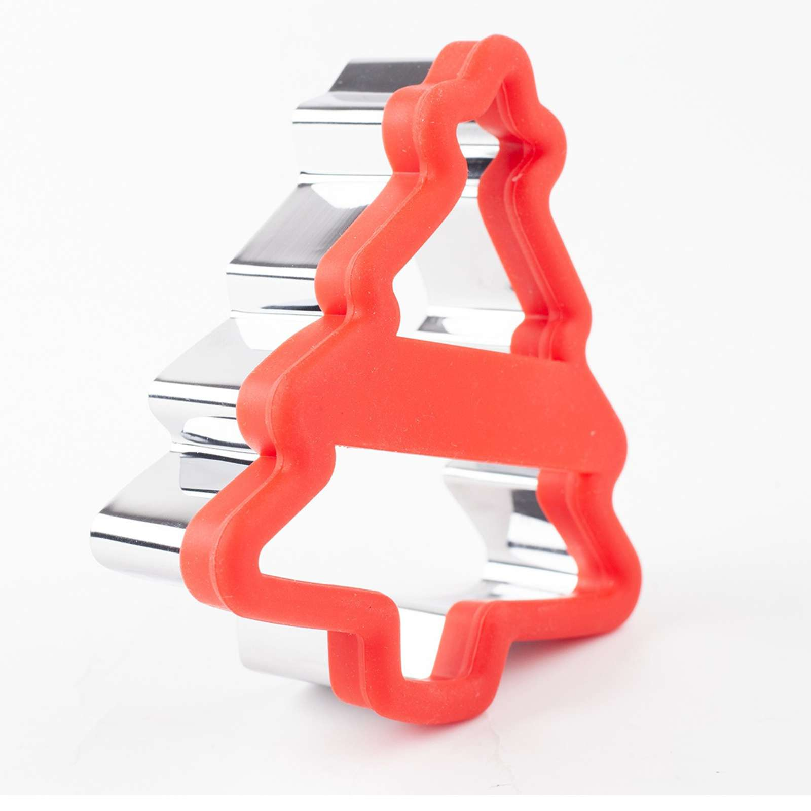 Ein roter, weihnachtsbaumförmiger Ausstecher liegt über einem silbernen, metallischen auf weißem Hintergrund.