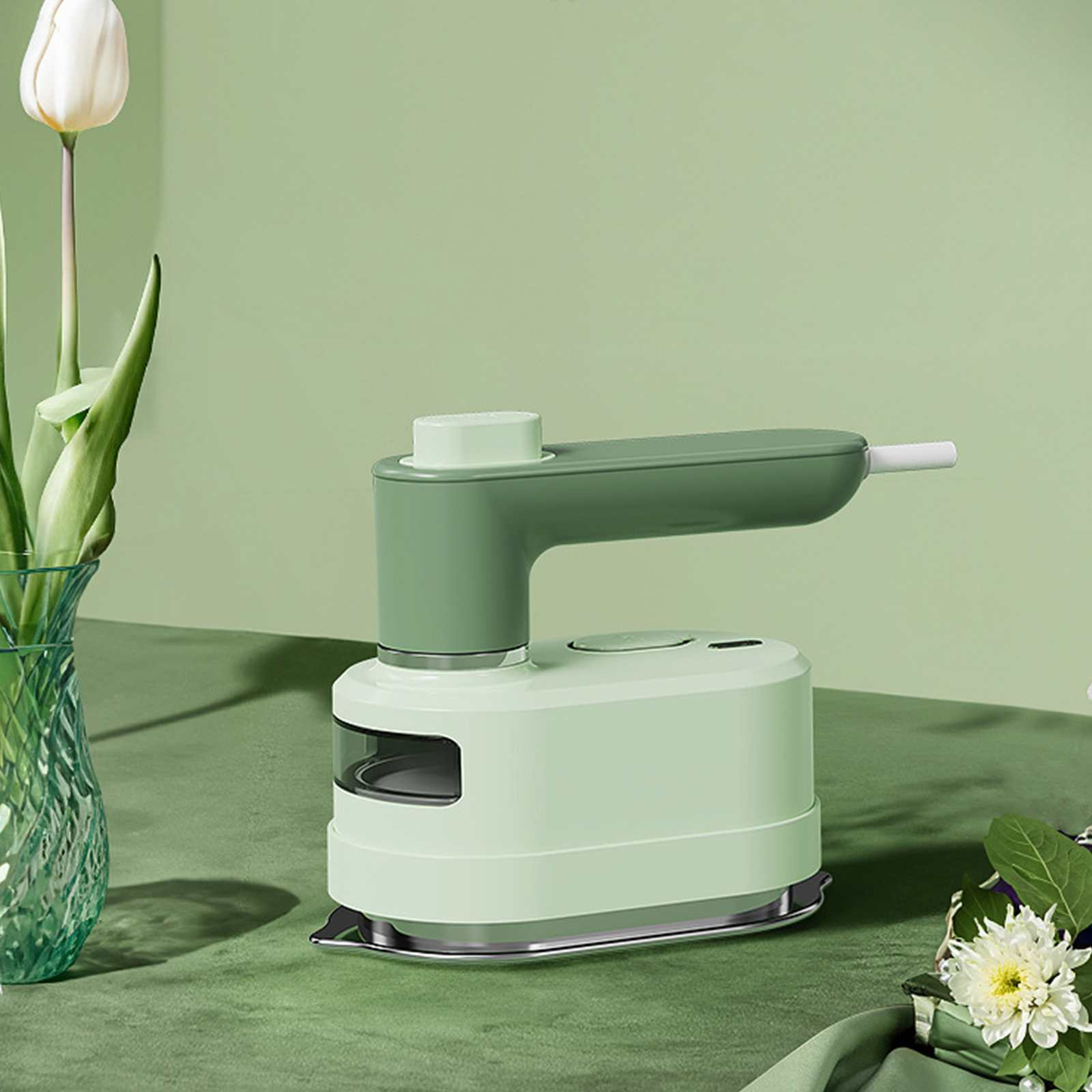 Ein mintgrünes und weißes Handbügeleisen auf einer grünen Oberfläche. Links eine Vase mit einer weißen Blume.