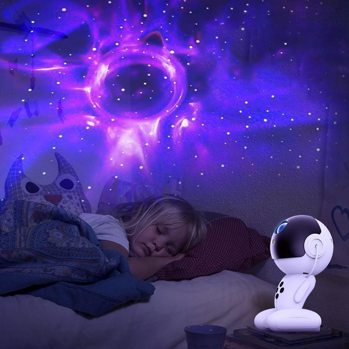 Ein Kind schläft unter einer Sternenhimmelprojektion. Ein weißes roboterartiges Nachtlicht steht daneben. Ein katzenförmiges Kissen liegt auf dem Bett.