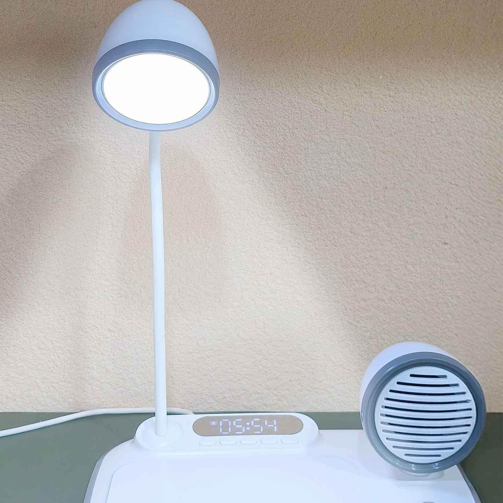 Eine weiße Schreibtischlampe mit rundem Lampenkopf steht auf einem weißen Sockel. Rechts ist ein Lautsprecher. Die Uhrzeit wird angezeigt.