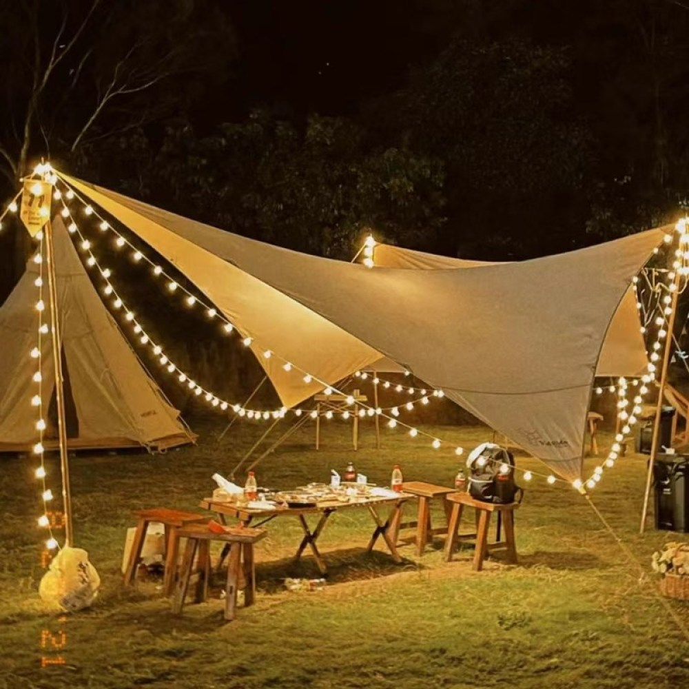 Eine nächtliche Campingszene mit Lichterketten, Zelten, Tischen und einem Vordach. Die Kulisse ist eine grasbewachsene Fläche mit Bäumen im Hintergrund.