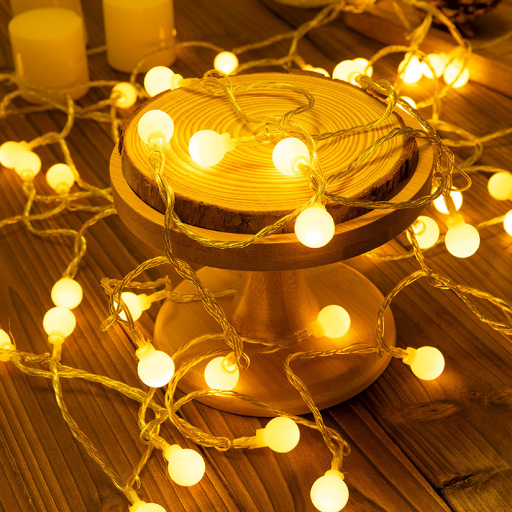 Ein hölzerner Kuchenständer mit einer Lichterkette aus warmweißen Kugellampen. Kerzen sind im Hintergrund. Die Szene spielt auf einer Holzoberfläche.