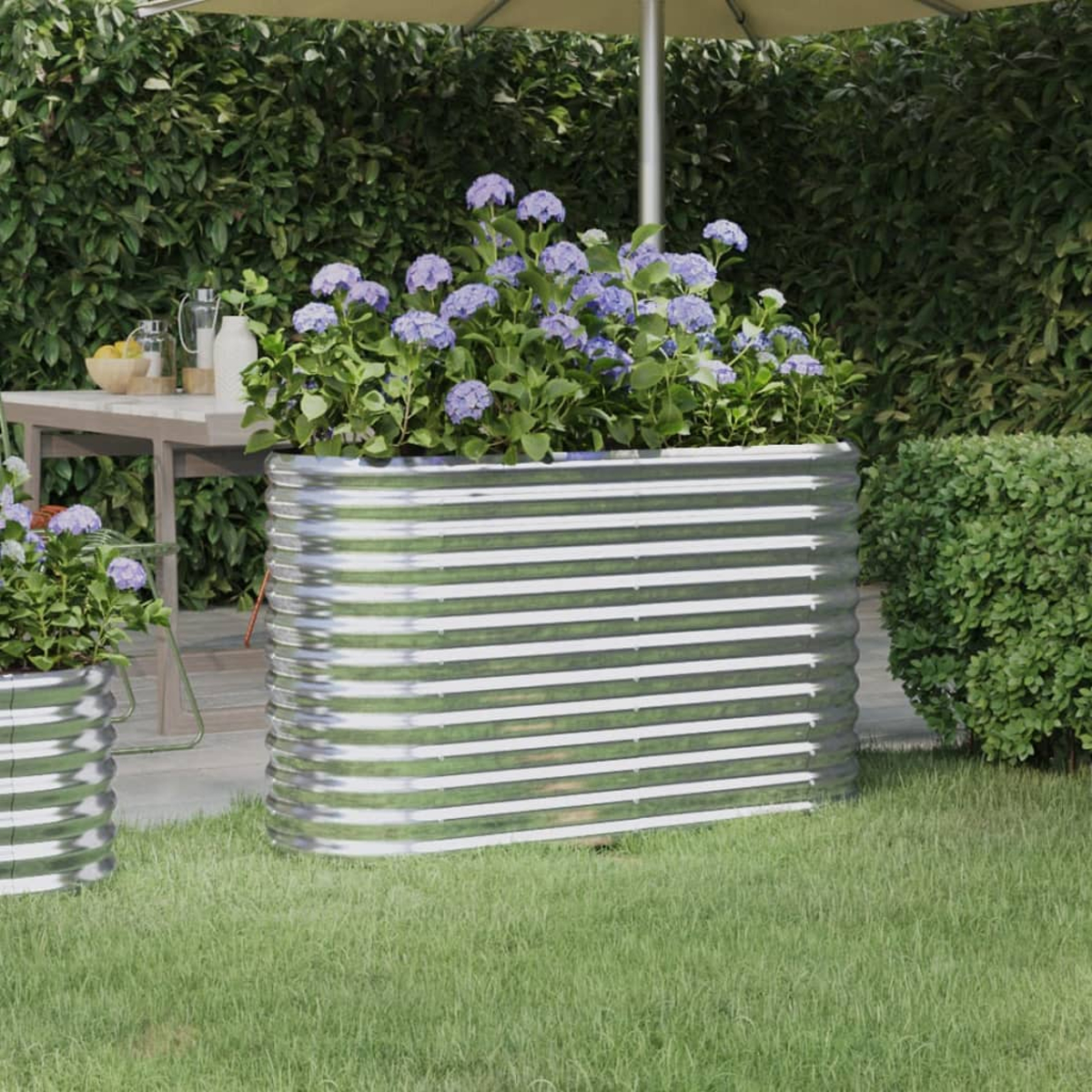 Metall-Pflanzgefäß mit blauen Blumen auf Gras. Tisch und Schirm im Hintergrund. Hecken bilden einen grünen Hintergrund.