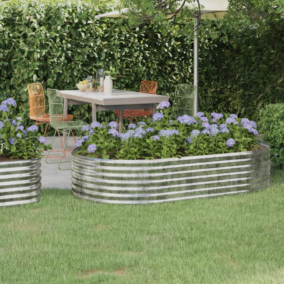 Gartenszene mit Blumen in Metallpflanzgefäßen, Tisch und Stühlen sowie grünem Laub. Ein Sonnenschirm spendet Schatten.