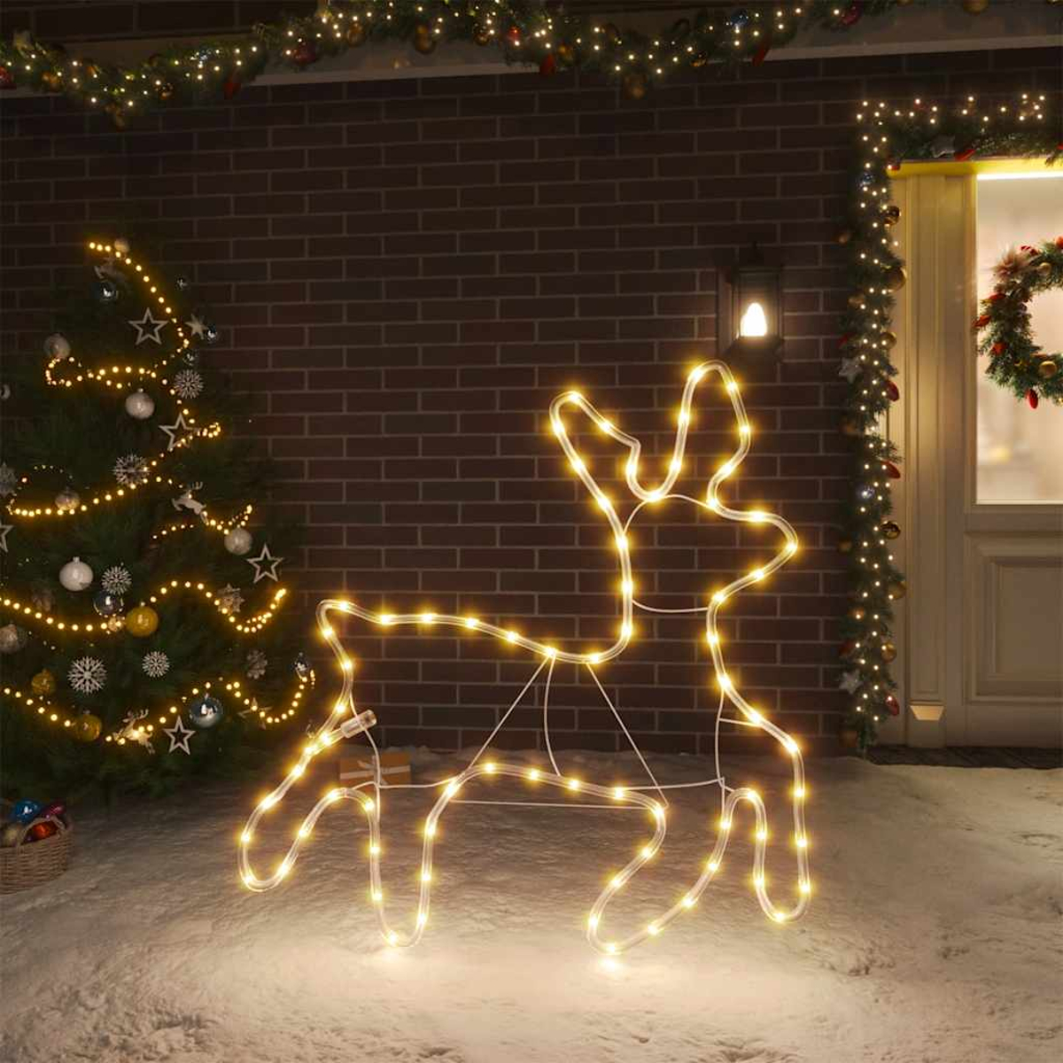 Eine leuchtende Rentierdekoration steht im Schnee. Ein Weihnachtsbaum und ein Hauseingang mit Lichtern und Girlanden sind im Hintergrund.