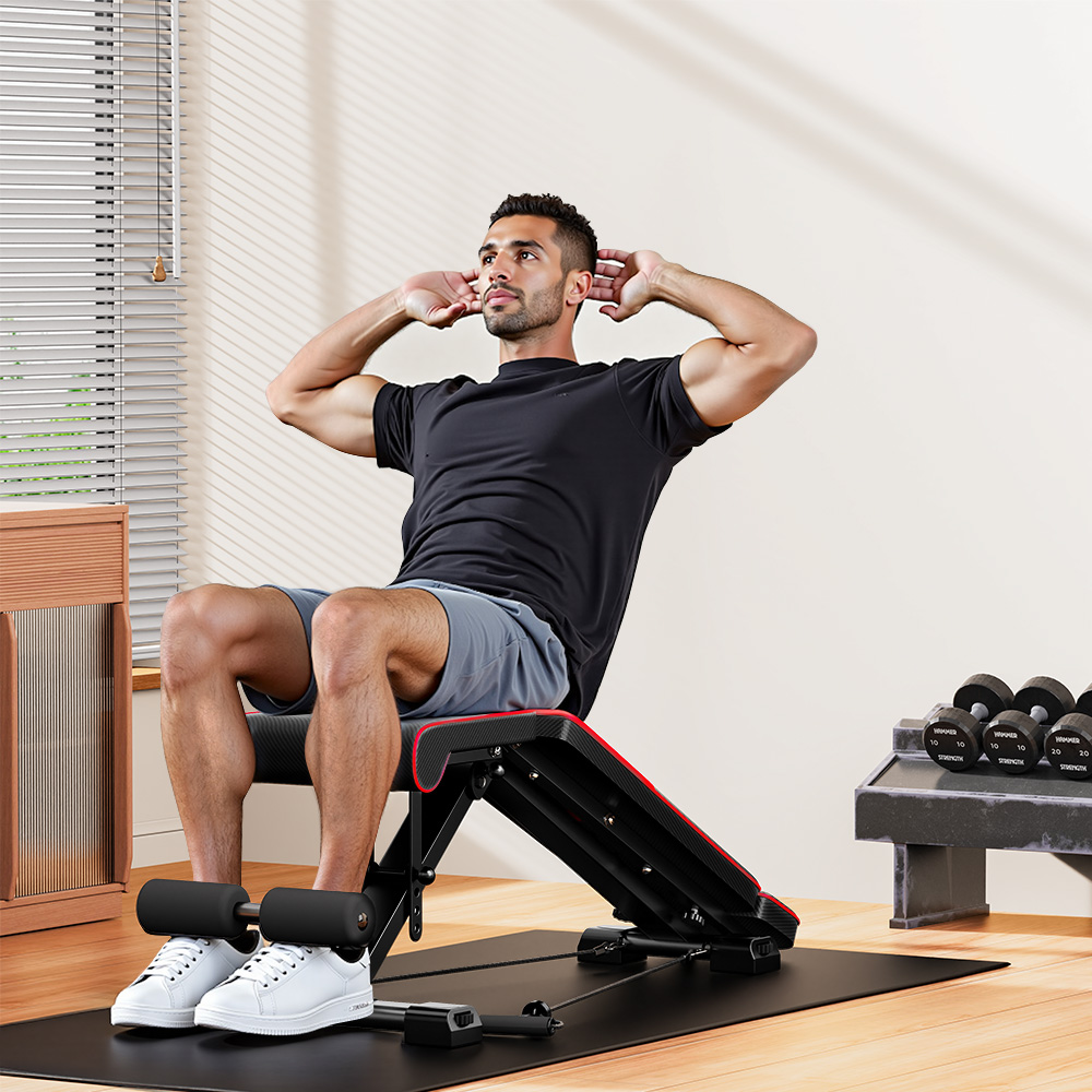 Mężczyzna robi brzuszki na ławce. Ma na sobie czarną koszulkę i szare szorty. Ławka ma czerwone wykończenie. Hantle są w tle.