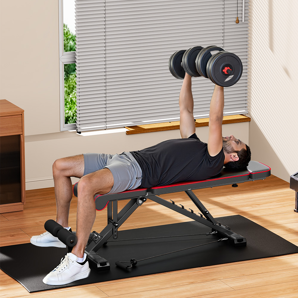 Mężczyzna leży na ławce i podnosi hantle. Jest w domowej siłowni. W tle okno z żaluzjami.