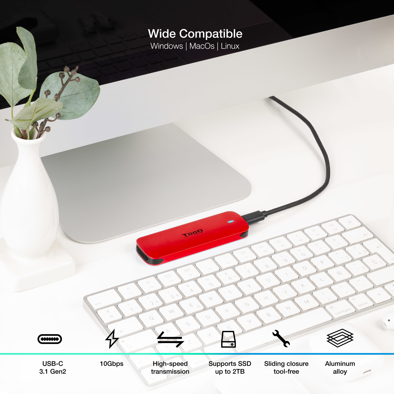 Un SSD externo rojo conectado a una computadora. Un jarrón blanco con vegetación a la izquierda y un teclado en primer plano.