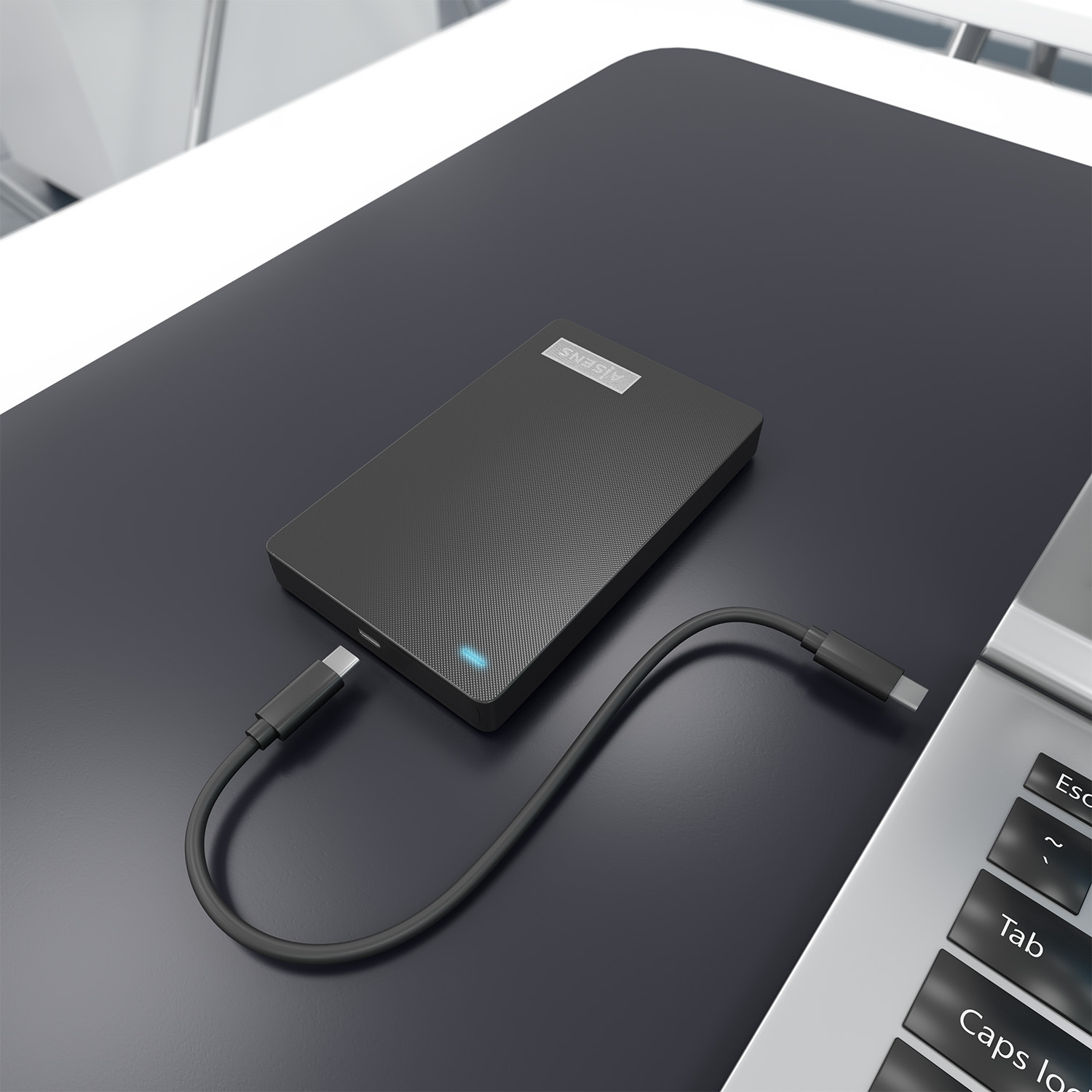Un disco duro externo negro sobre una superficie negra, conectado por un cable USB-C a un portátil. El teclado del portátil es visible.