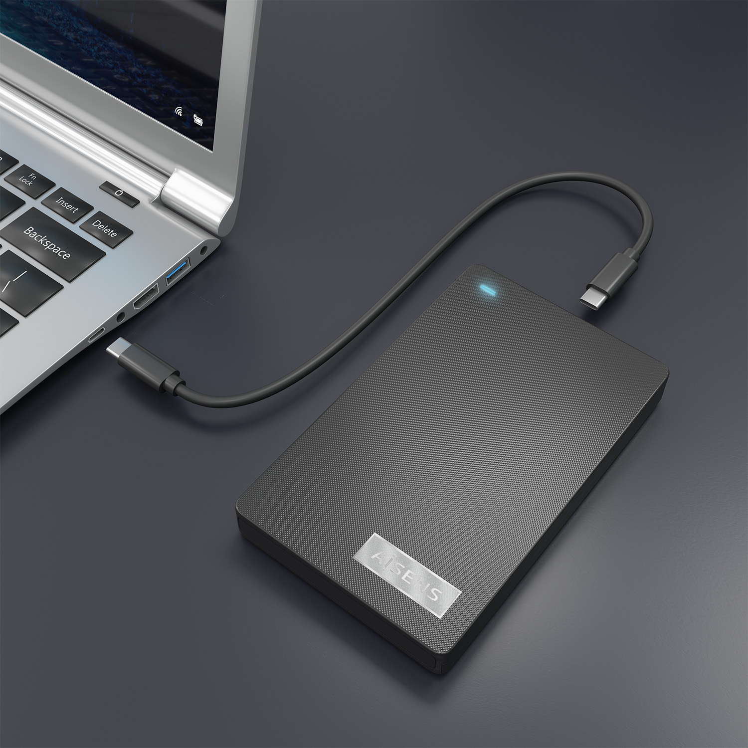 Un disco duro externo negro está conectado a un portátil con un cable USB-C. El portátil es plateado y el fondo es gris oscuro.