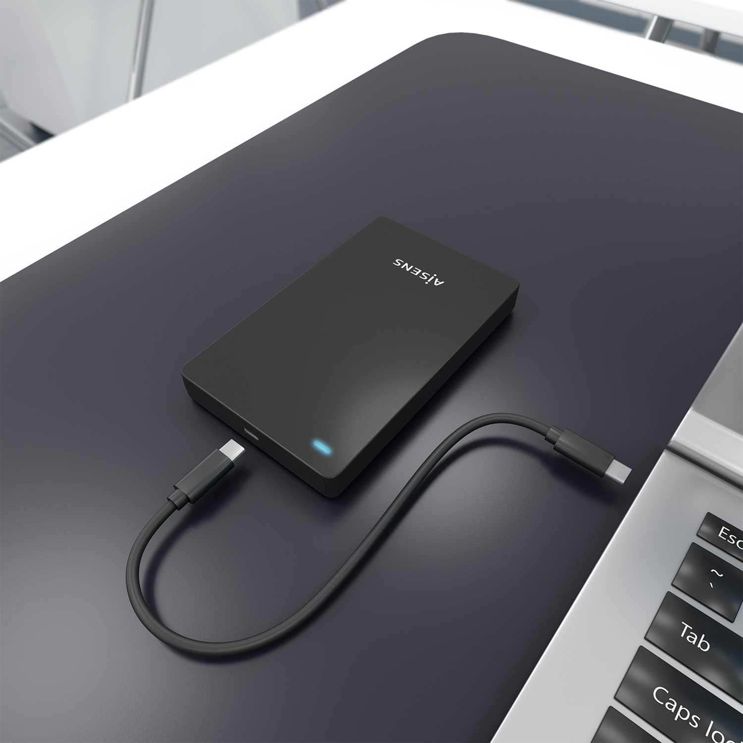 Un disco duro externo negro con un cable USB-C conectado sobre una superficie oscura. Parte del teclado de un portátil es visible.