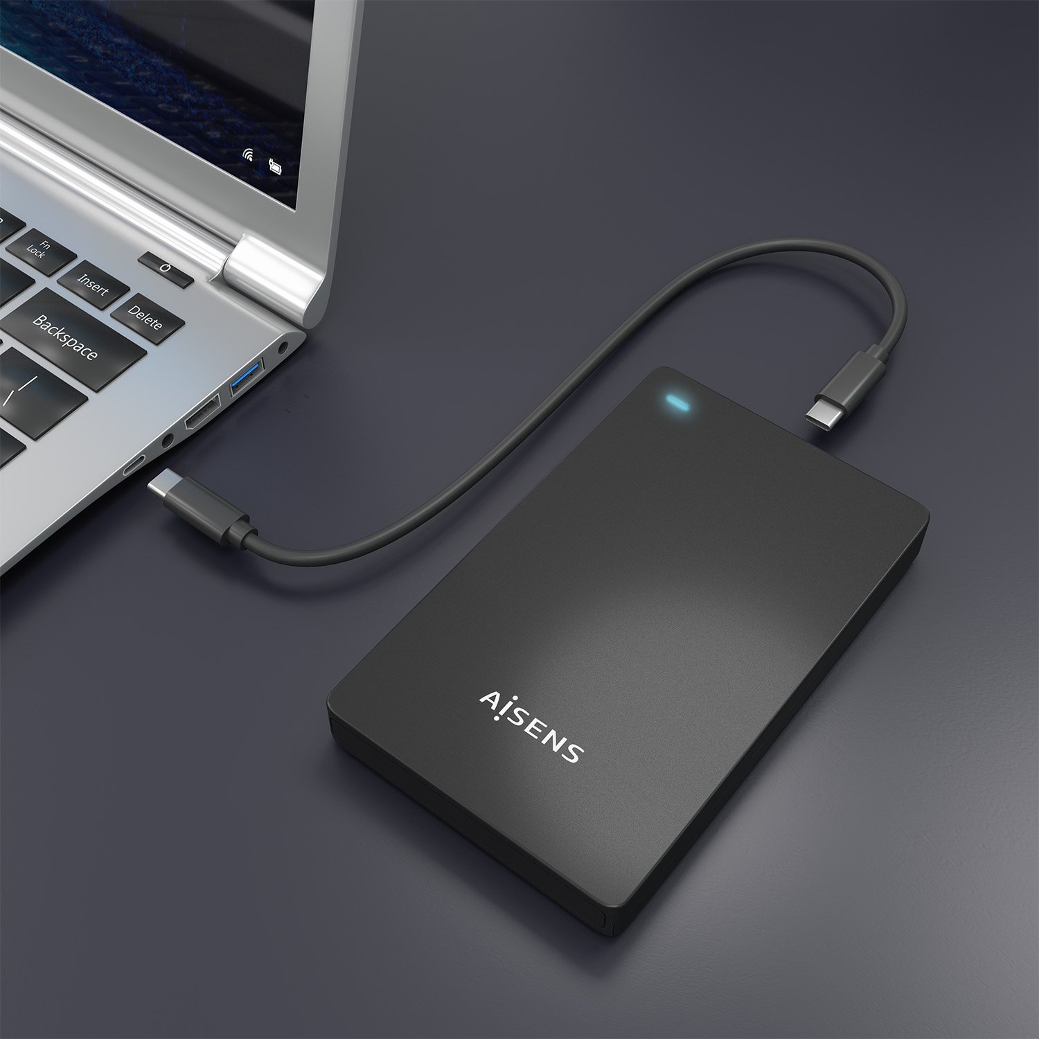 Un disco duro externo negro conectado a una laptop a través de un cable USB-C, ambos sobre una superficie gris oscura.