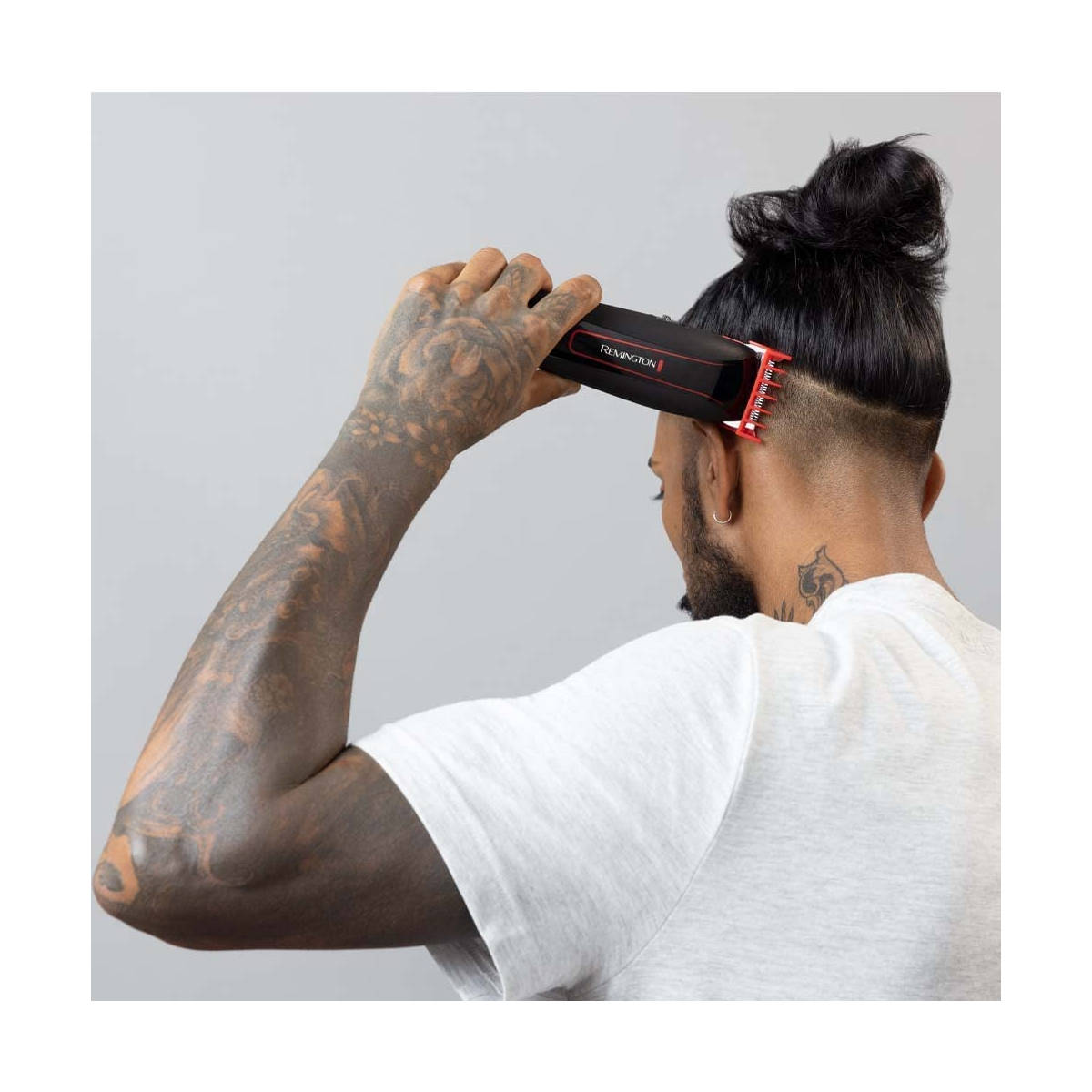 Un uomo con tatuaggi sul braccio usa un tagliacapelli Remington. Ha uno chignon e la nuca rasata. Indossa una maglietta bianca.