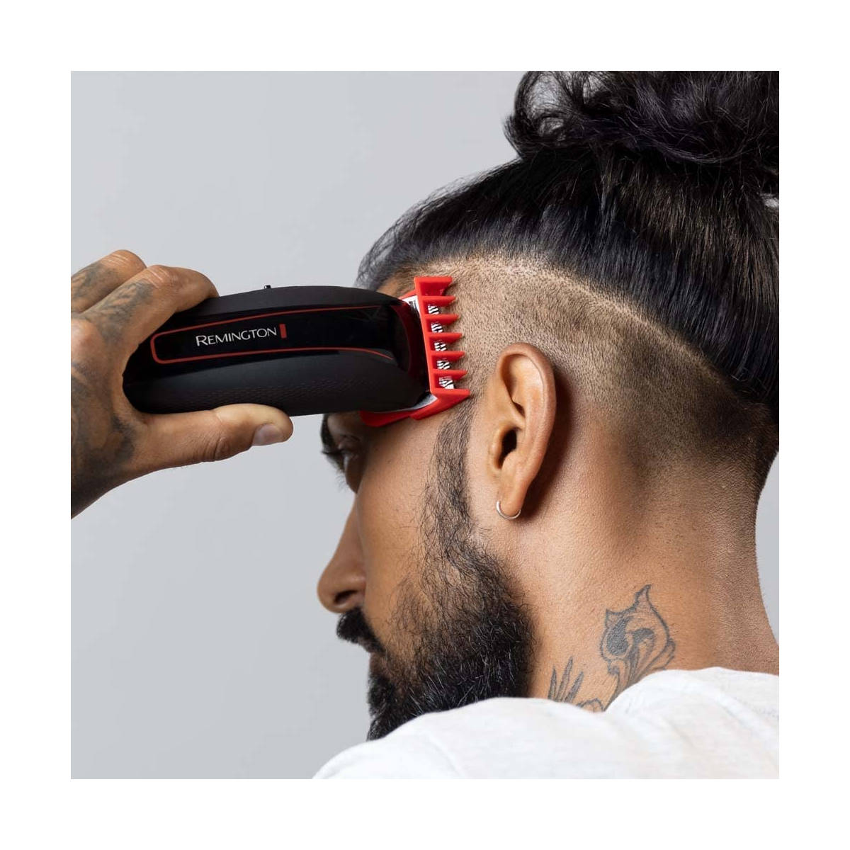 Un uomo si rade i capelli con un tagliacapelli Remington. Ha la barba, un tatuaggio e uno chignon. Il tagliacapelli è nero e rosso.