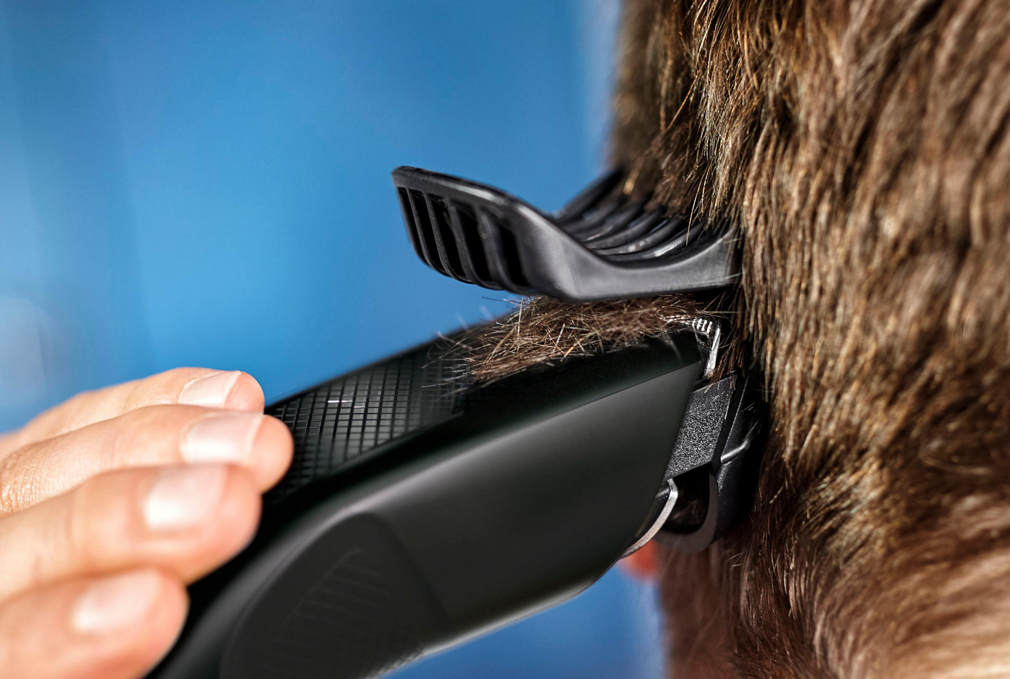 Ein schwarzer Haarschneider schneidet braunes Haar. Eine Hand hält den Haarschneider. Der Hintergrund ist blau.