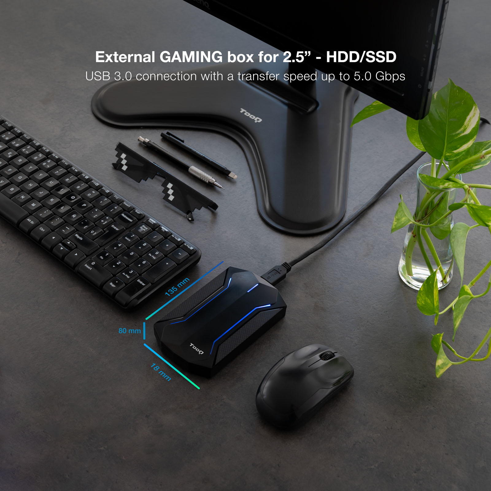 Una caja de juegos externa, teclado negro, ratón, bolígrafos y una planta en un escritorio. También se muestran las dimensiones.