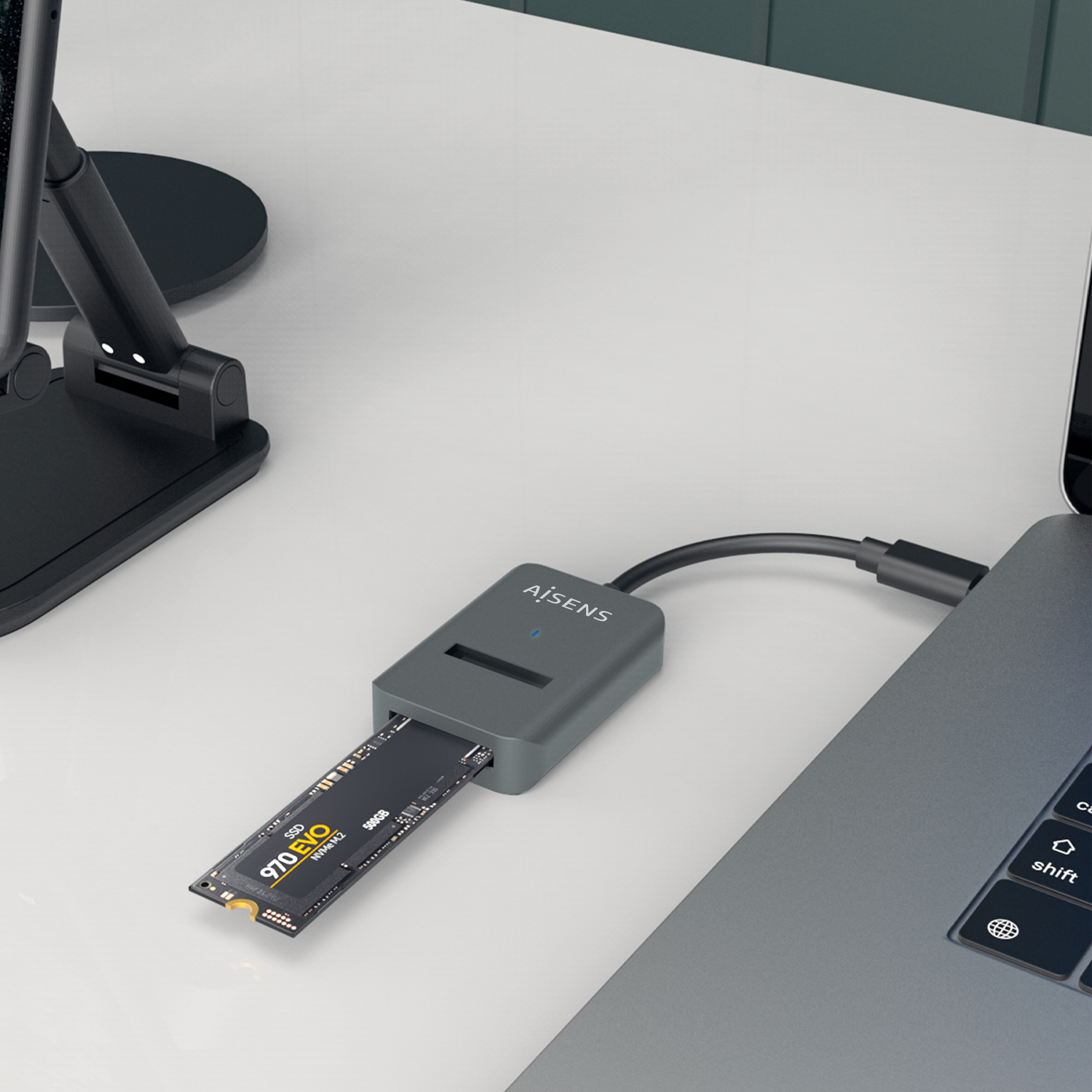 Un SSD externo gris conectado a una laptop. El SSD está etiquetado como '970 EVO' y '500GB'. A la izquierda hay un soporte para teléfono negro.