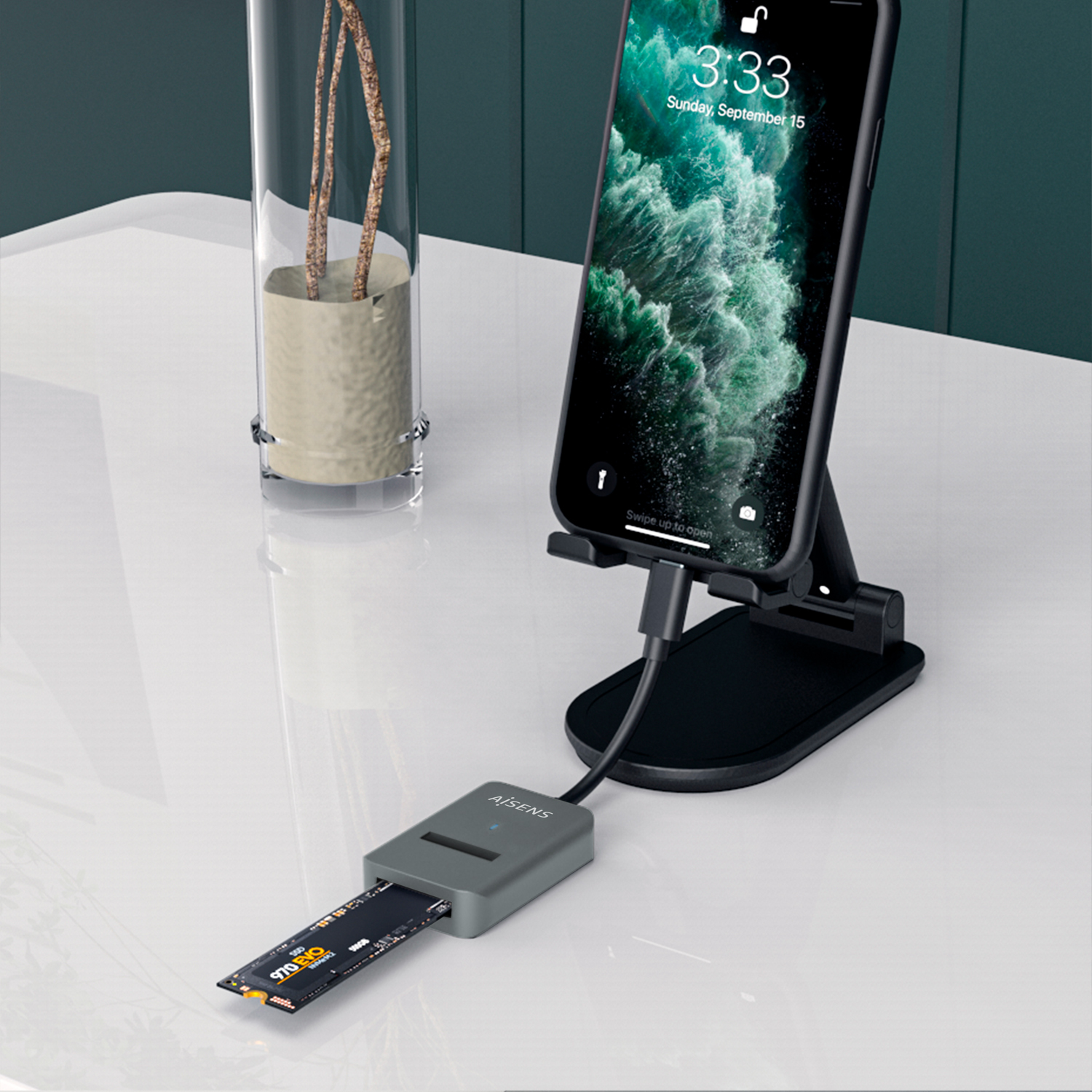 Un teléfono en un soporte, conectado a un lector de tarjetas gris, y un jarrón con ramas. El teléfono muestra la hora.