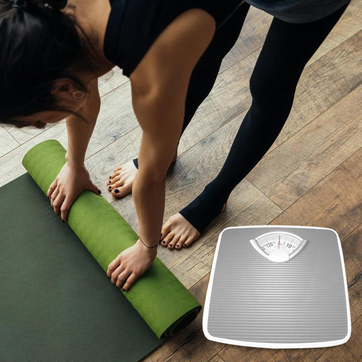 Eine Frau rollt eine grüne Yogamatte aus, im Hintergrund eine graue Waage. Sie trägt eine schwarze Leggings und ein dunkles Oberteil.