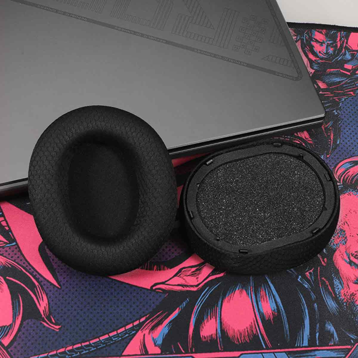Zwei schwarze Kopfhörer-Ohrmuscheln auf einem Gaming-Mauspad und einem Laptop. Eine Ohrmuschel ist abgenommen.