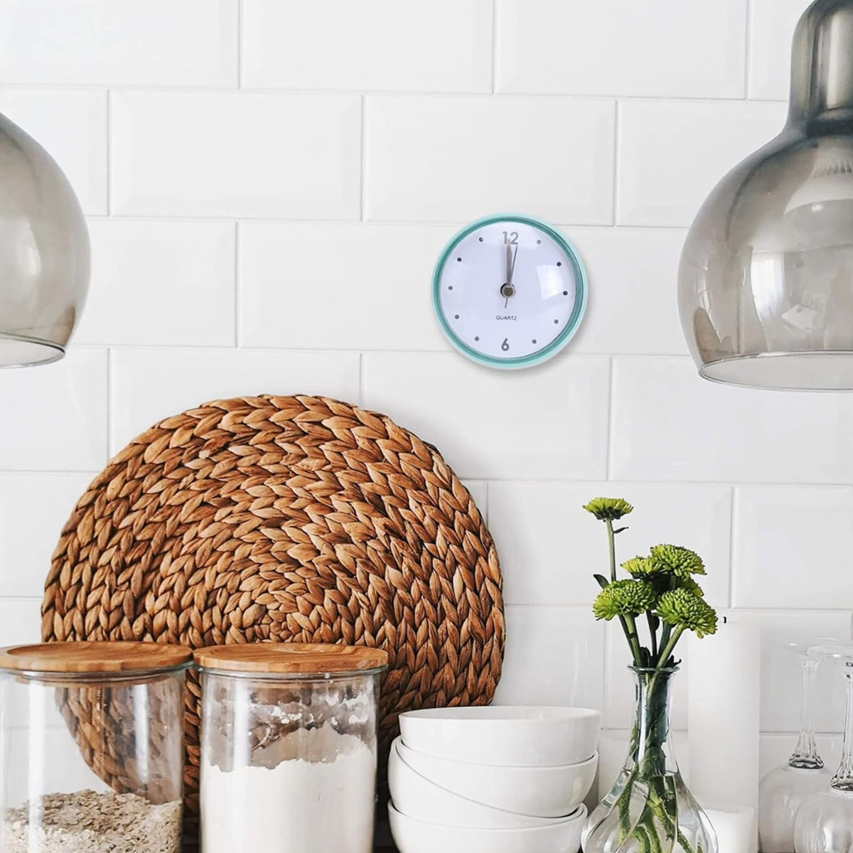 Küchenszene mit Uhr, gewebter Matte, Gläsern, Schüsseln und Blumen. Die Uhr zeigt 12 Uhr. Weiß geflieste Wand im Hintergrund.