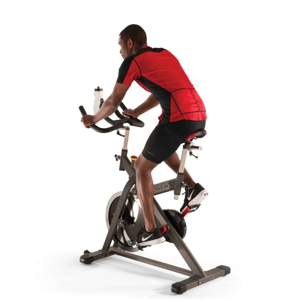 Un homme fait du vélo d'exercice stationnaire. Il porte une chemise rouge et noire et un short noir. Le vélo est gris, avec une bouteille d'eau.