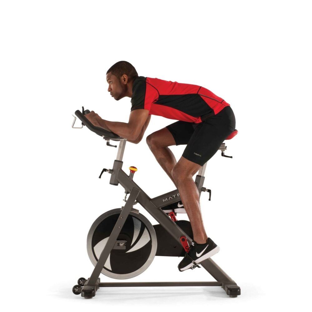 Un homme en tenue de sport fait du vélo d'appartement. Il porte un short noir, une chemise rouge et noire et des baskets noires.