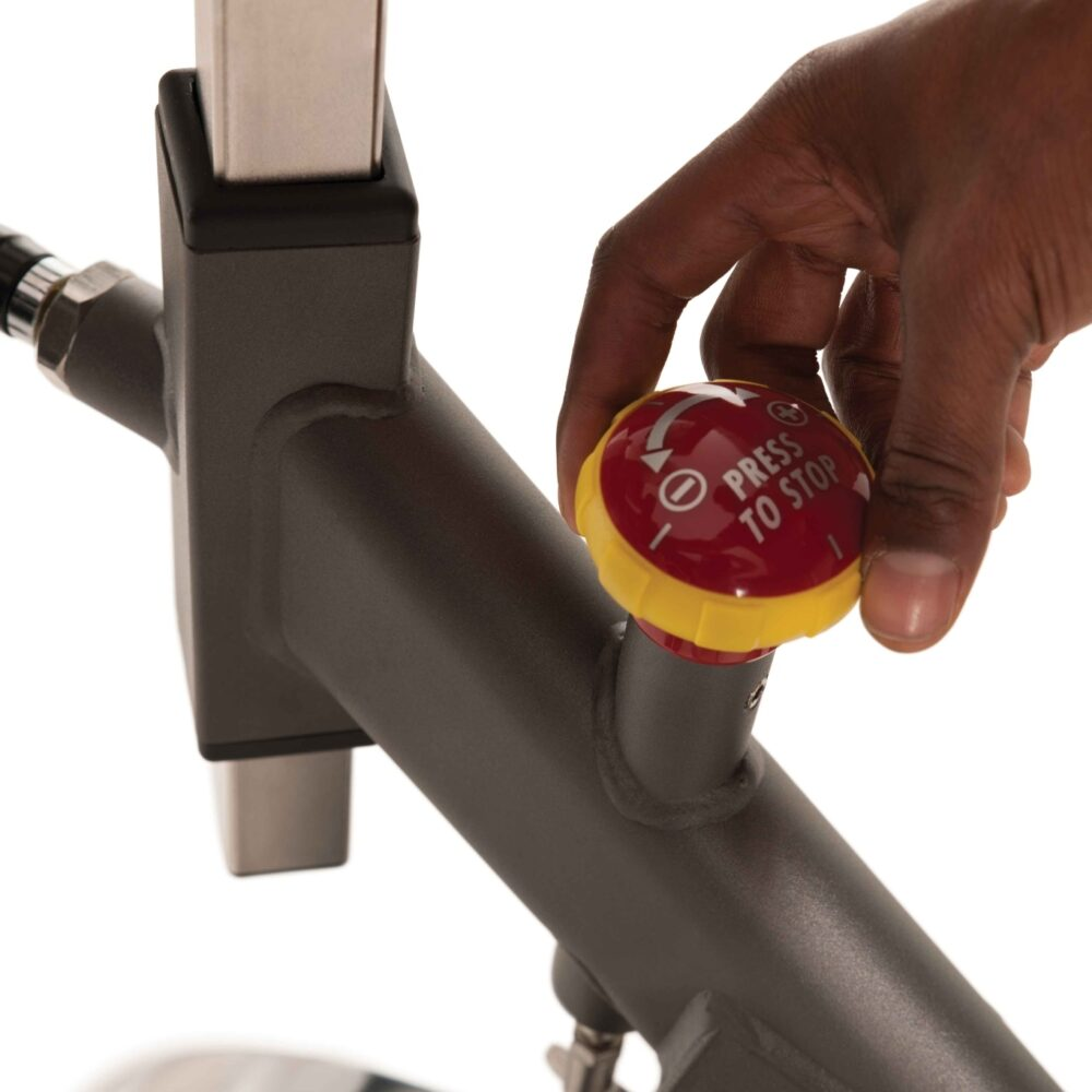 Main tournant un bouton rouge et jaune avec l'inscription 'PRESS TO STOP' sur un vélo d'appartement. Le cadre du vélo est gris.