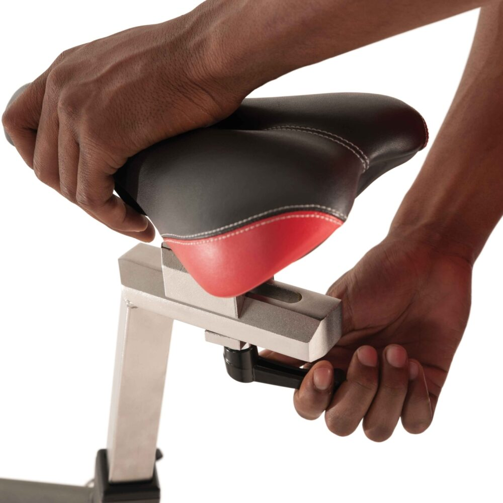 Personne ajustant la selle d'un vélo d'appartement. Selle noire et rouge avec coutures blanches. Mains sur la selle et le levier de réglage.