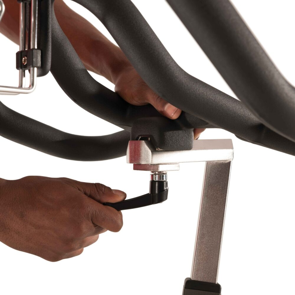 Mains ajustant un levier noir sur un vélo d'exercice argenté et noir. Les mains sont de peau foncée. Focus sur les pièces mécaniques.