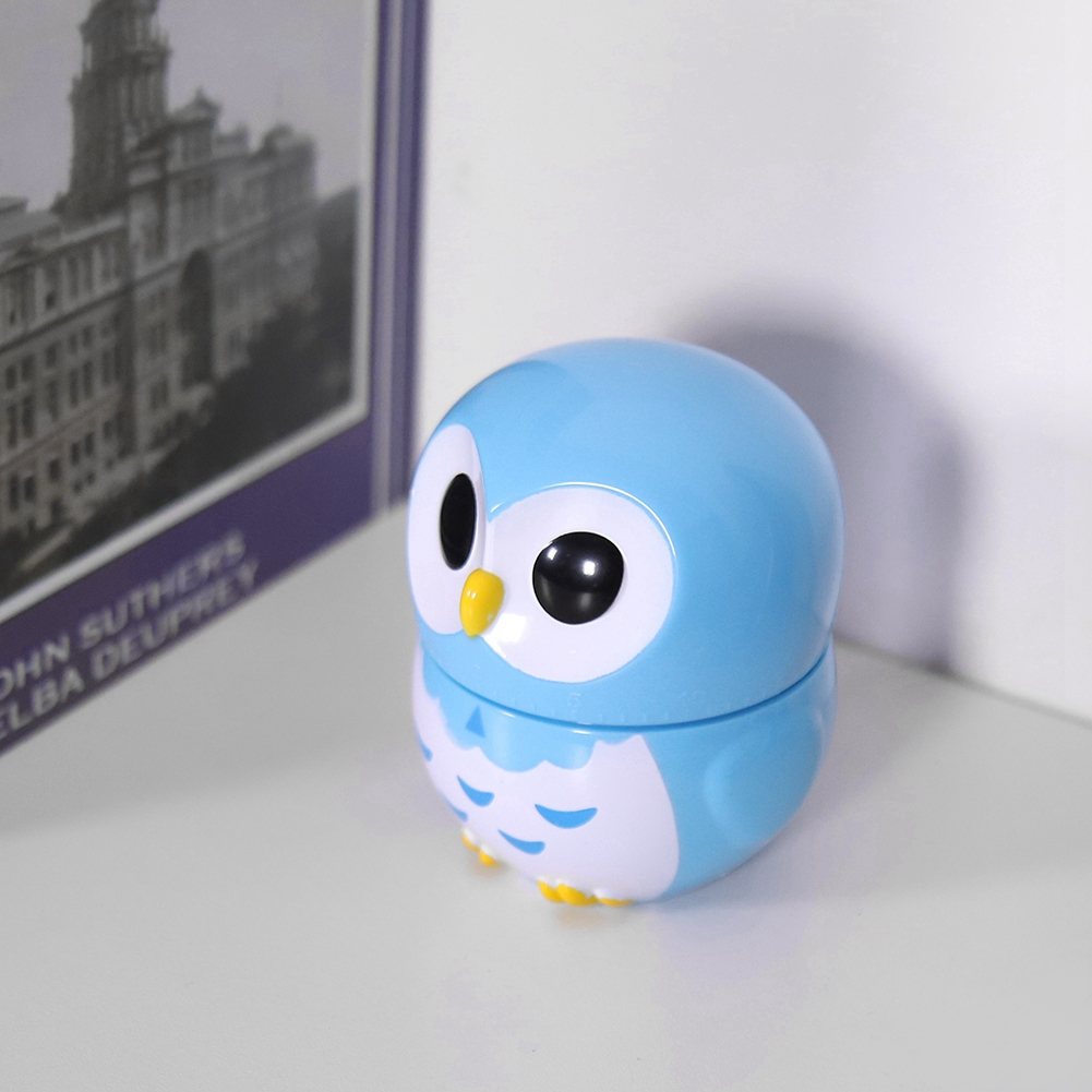Ein blaues eulenformiges Behältnis steht auf einer weißen Oberfläche. Im Hintergrund befindet sich ein Buch. Die Eule hat große schwarze Augen und einen gelben Schnabel.
